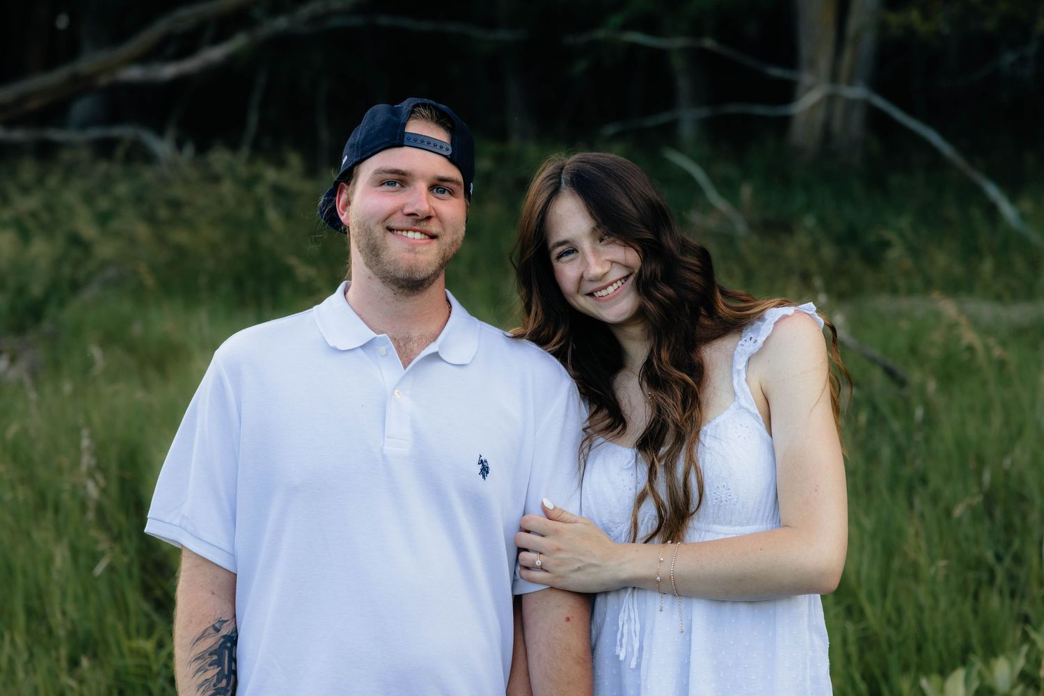 Happy engaged couple in white outfits smiling together in a lush green meadow setting.