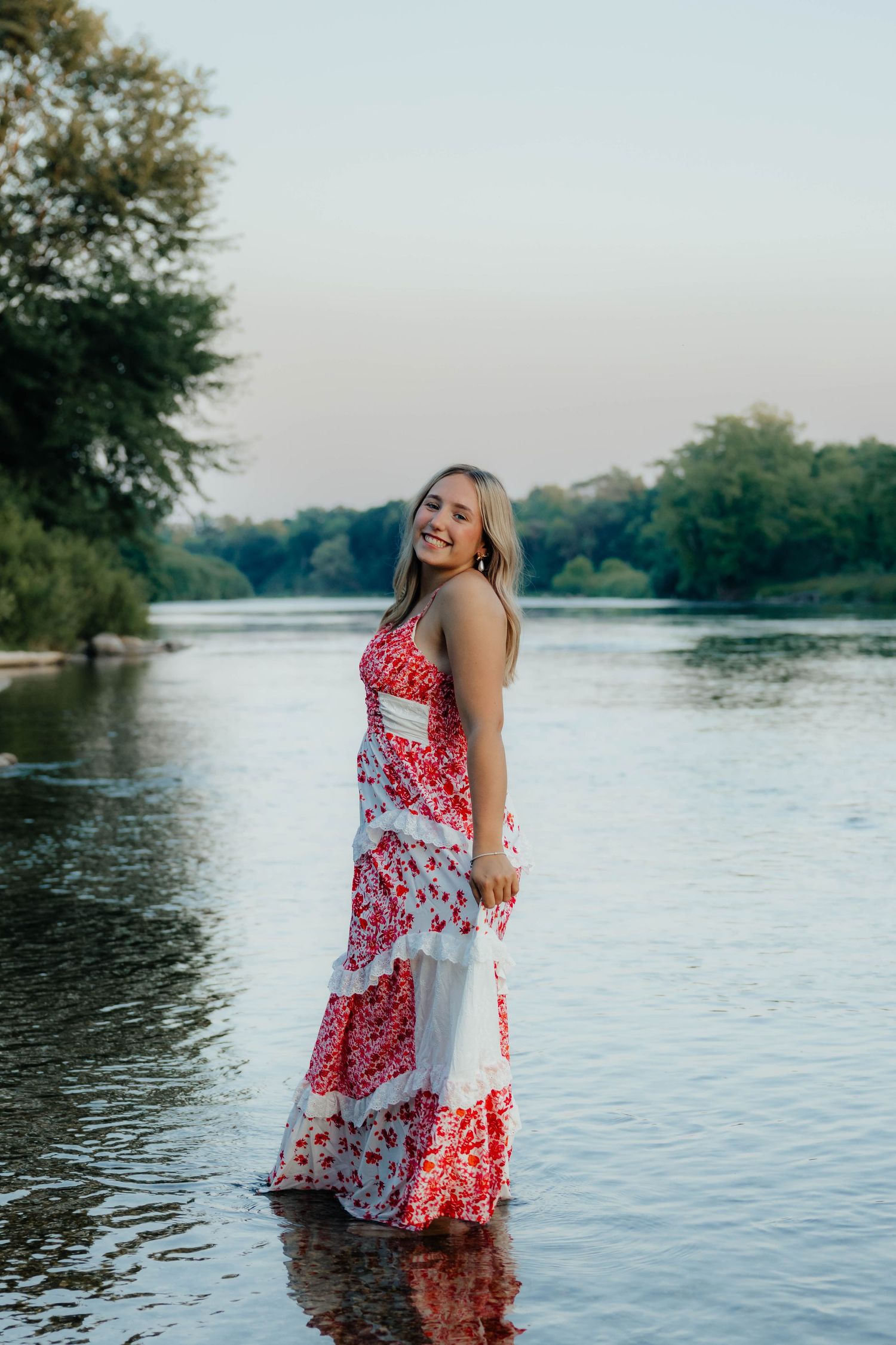 Blonde woman in red and white floral maxi dress standing in shallow river at dusk smiling.