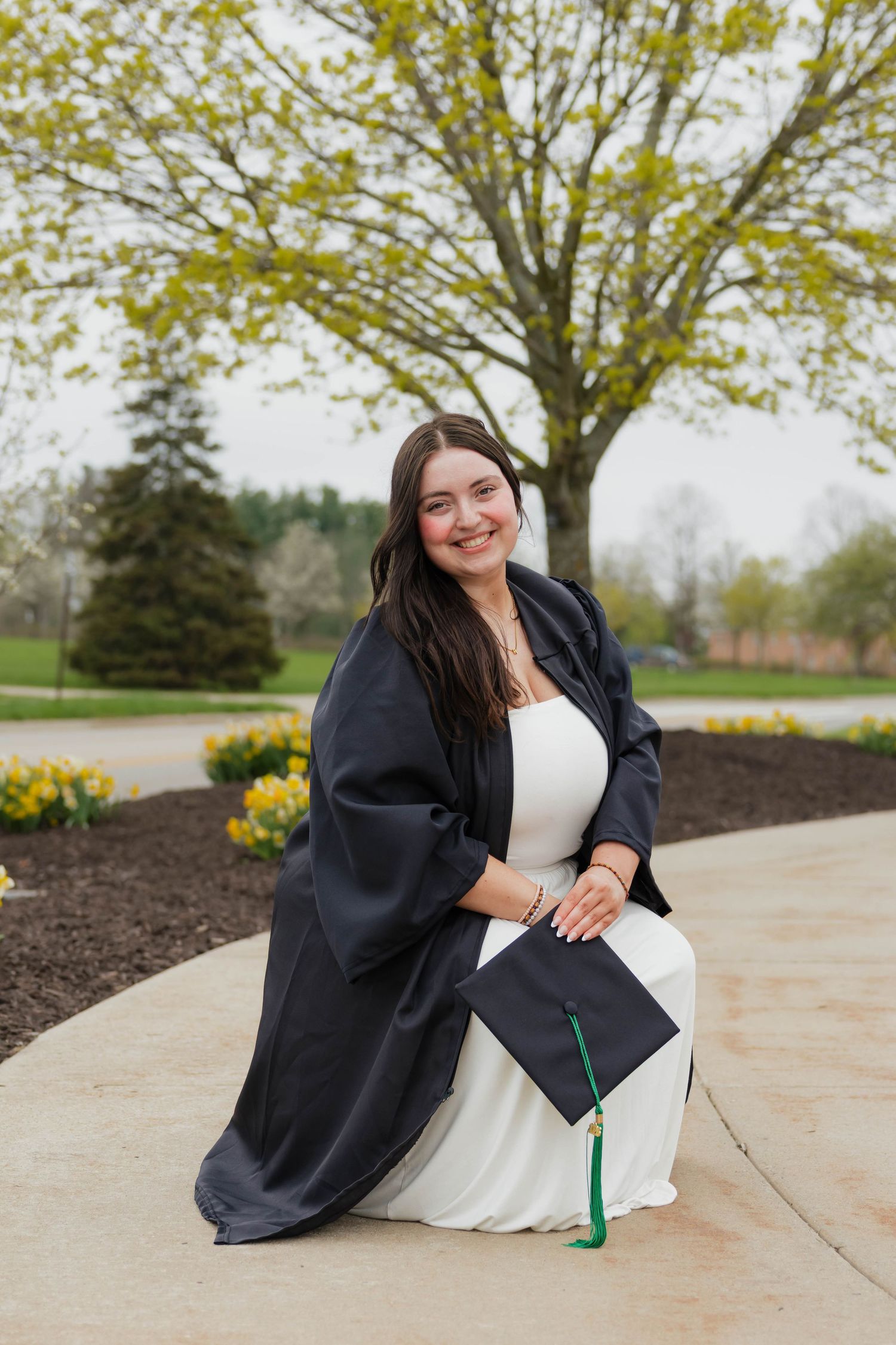 A smiling graduate in a white dress and black gown kneels outdoors holding her green-tasseled cap.