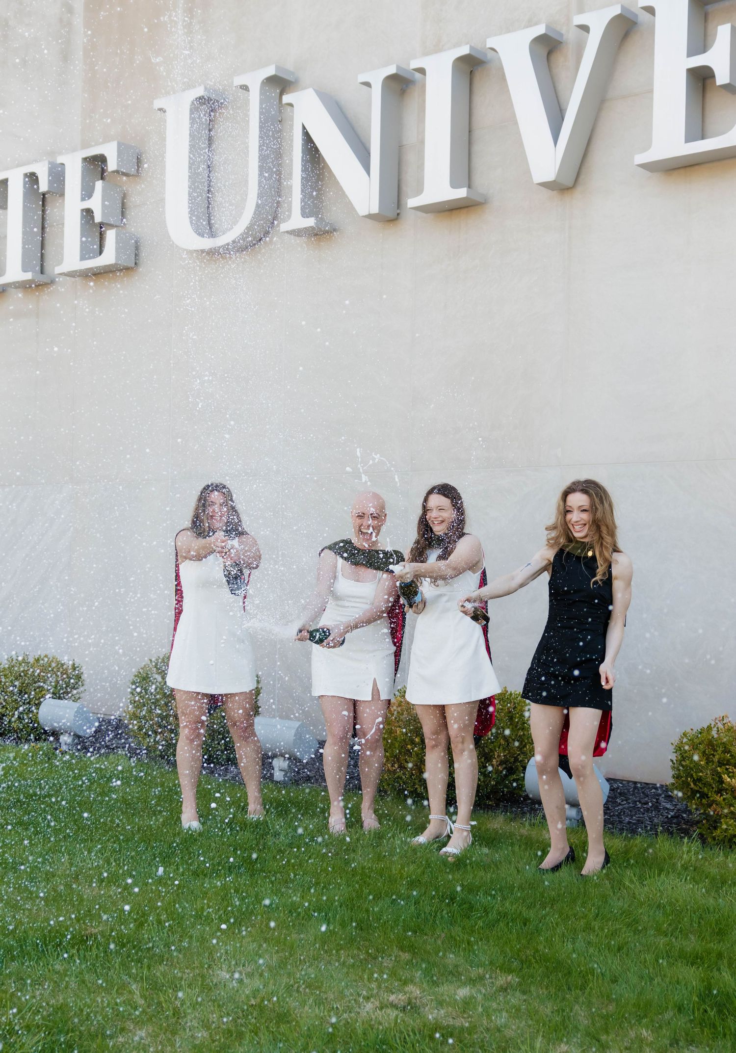 Four women in white and black dresses celebrate by popping champagne bottles at a university sign.