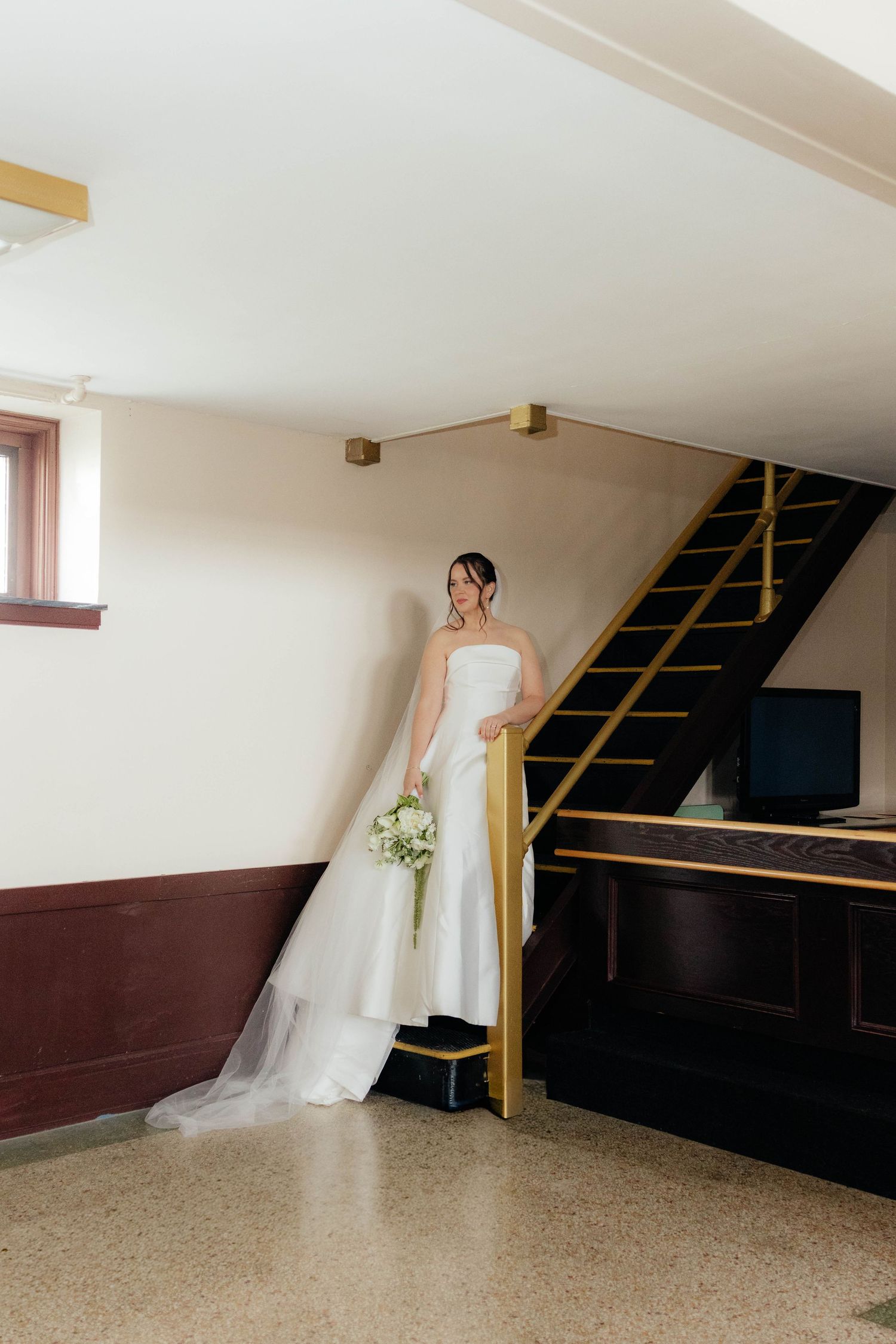Bride in minimalist strapless wedding gown standing by gold staircase railing in vintage venue.