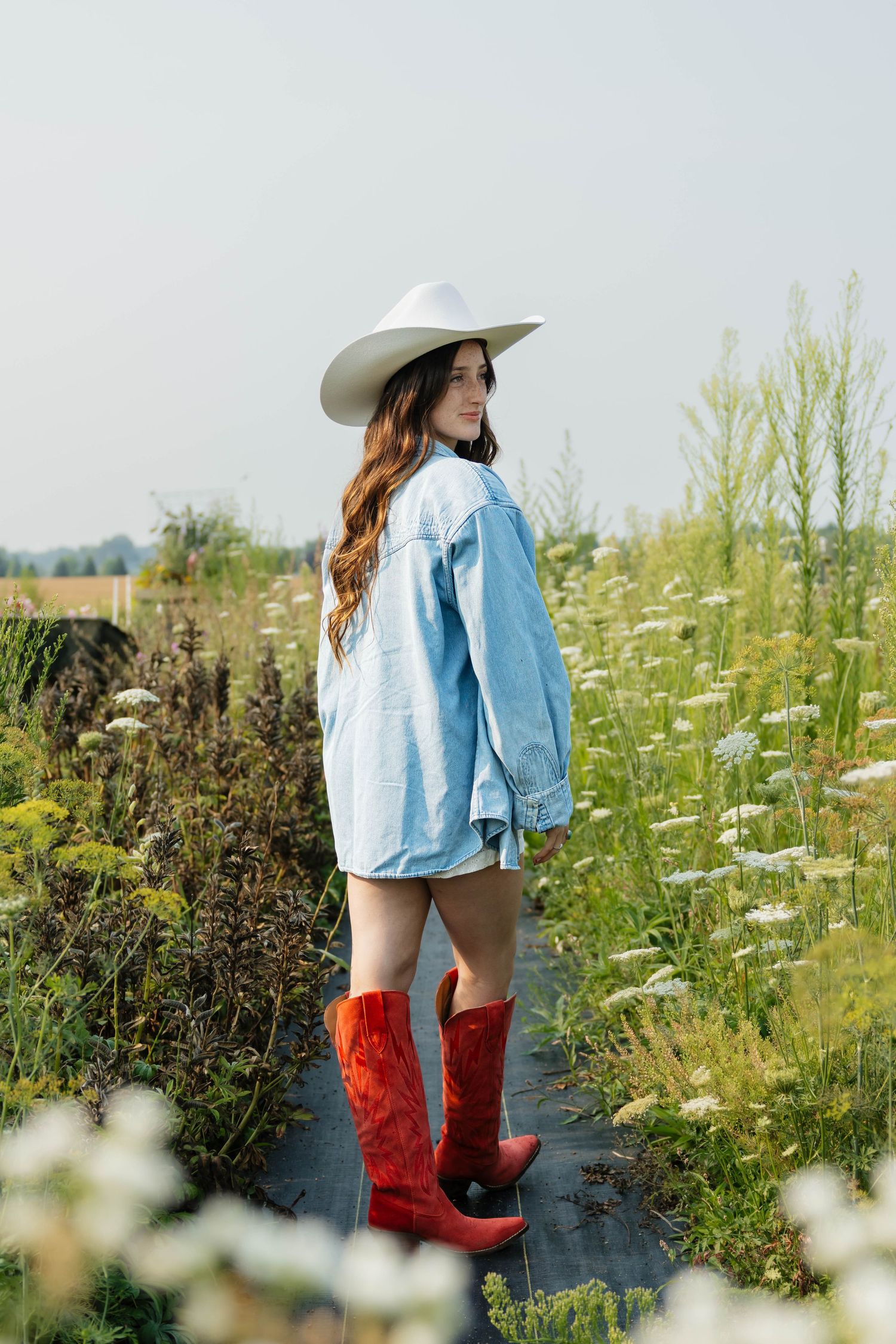 Young woman in white cowboy hat, denim jacket, and red cowboy boots standing in a wildflower field.