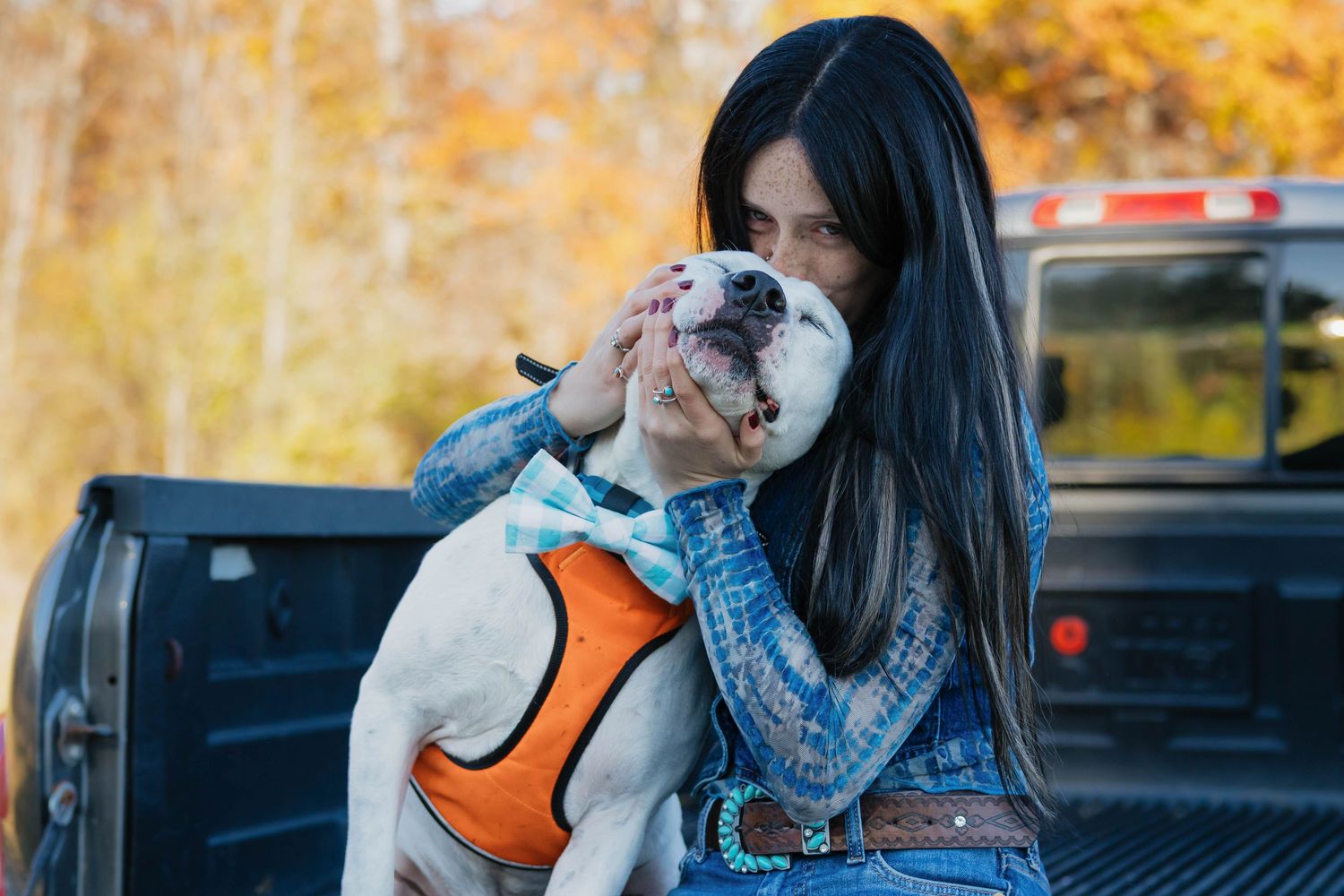 Woman with long dark hair kissing a white pitbull dog wearing an orange harness on a truck bed.