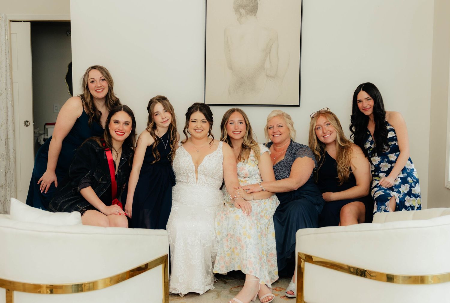 Bride in white gown poses with seven bridesmaids and guests on white sofa during wedding celebration.
