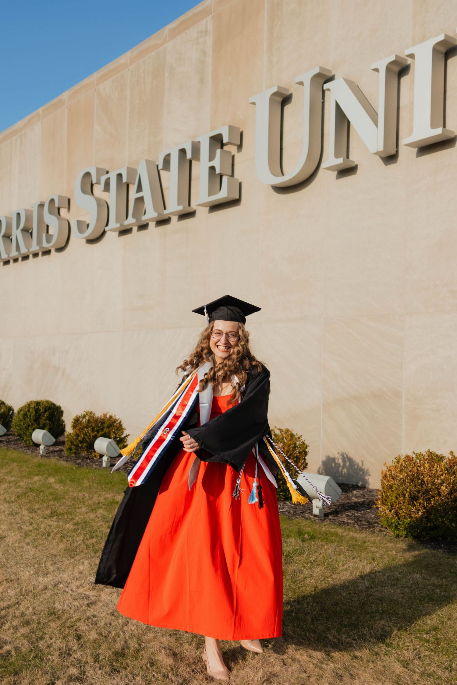 Smiling graduate in orange dress and black gown poses with colorful stoles in front of Ferris State University sign.