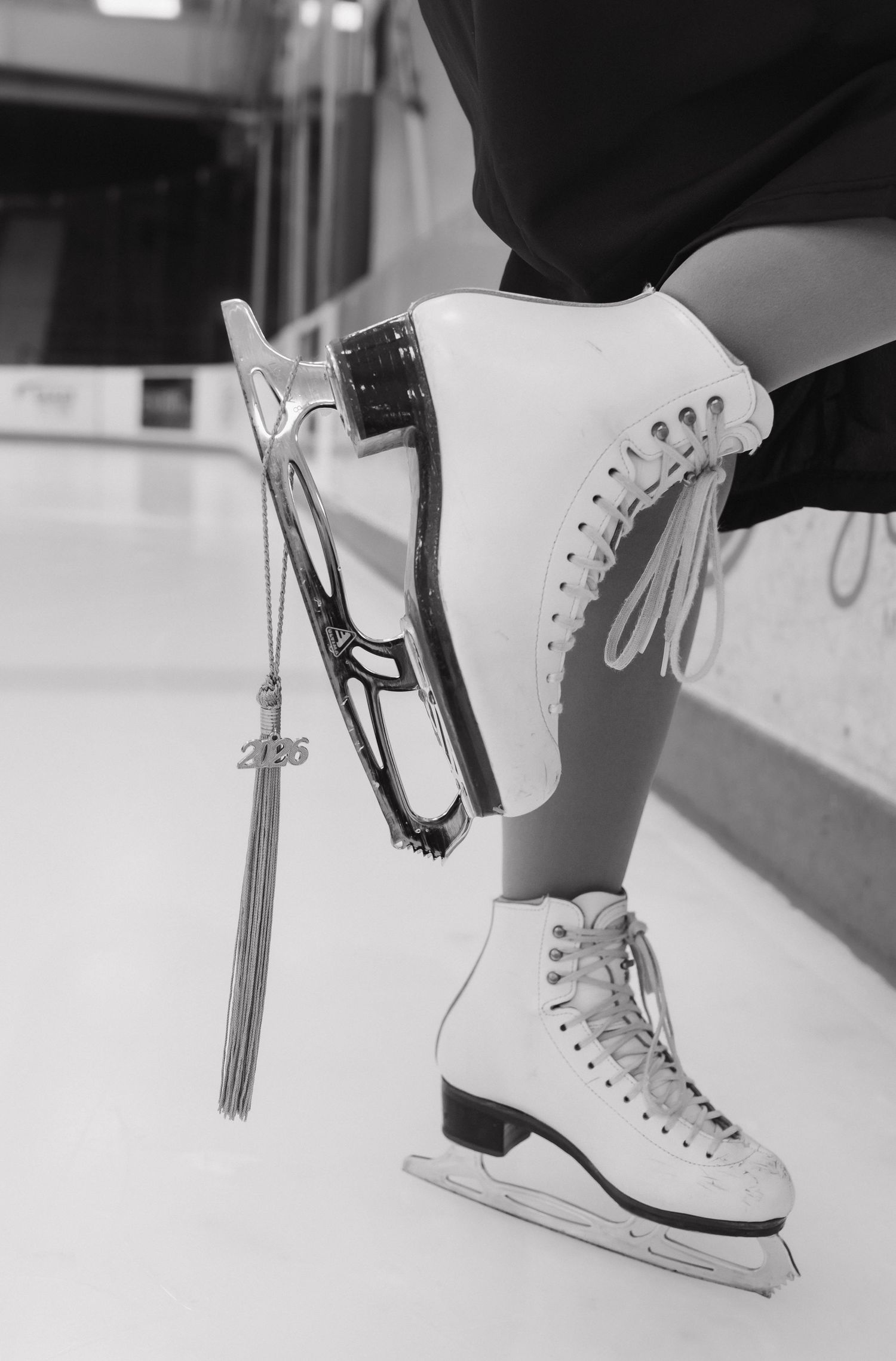 Black and white close-up of white figure ice skates with graduation tassel charm at rink.