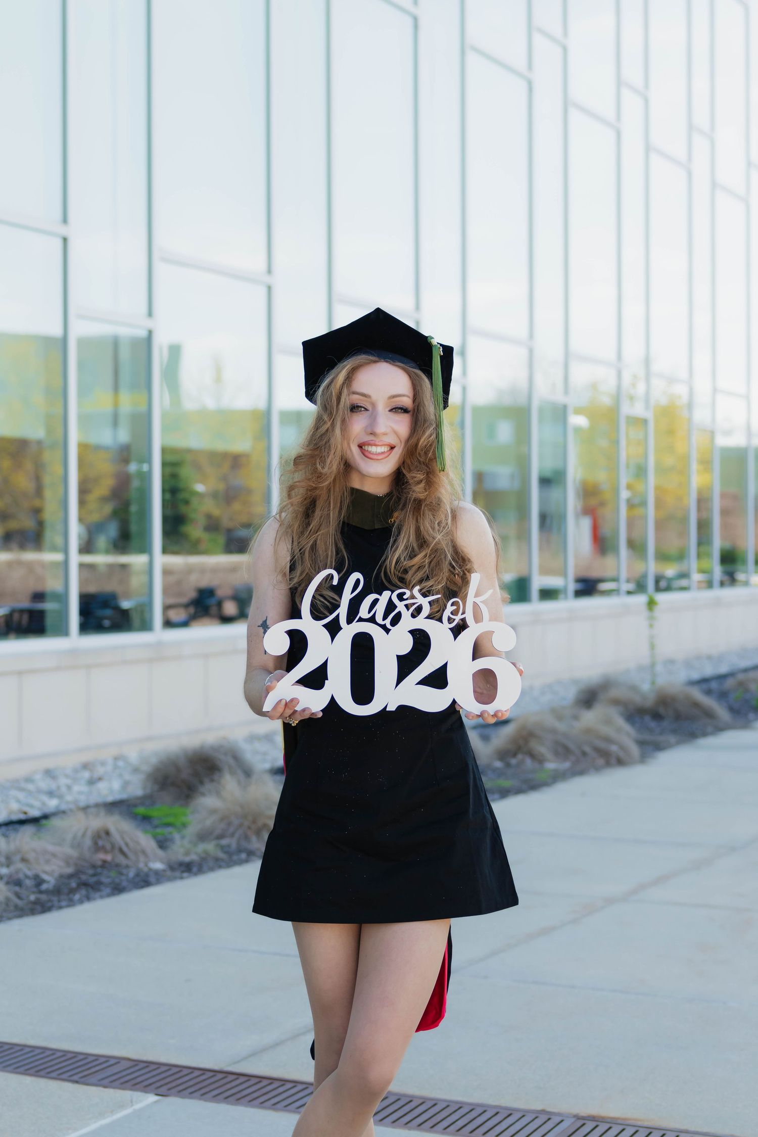 Smiling blonde graduate in black dress and cap holds Class of 2026 sign outdoors.