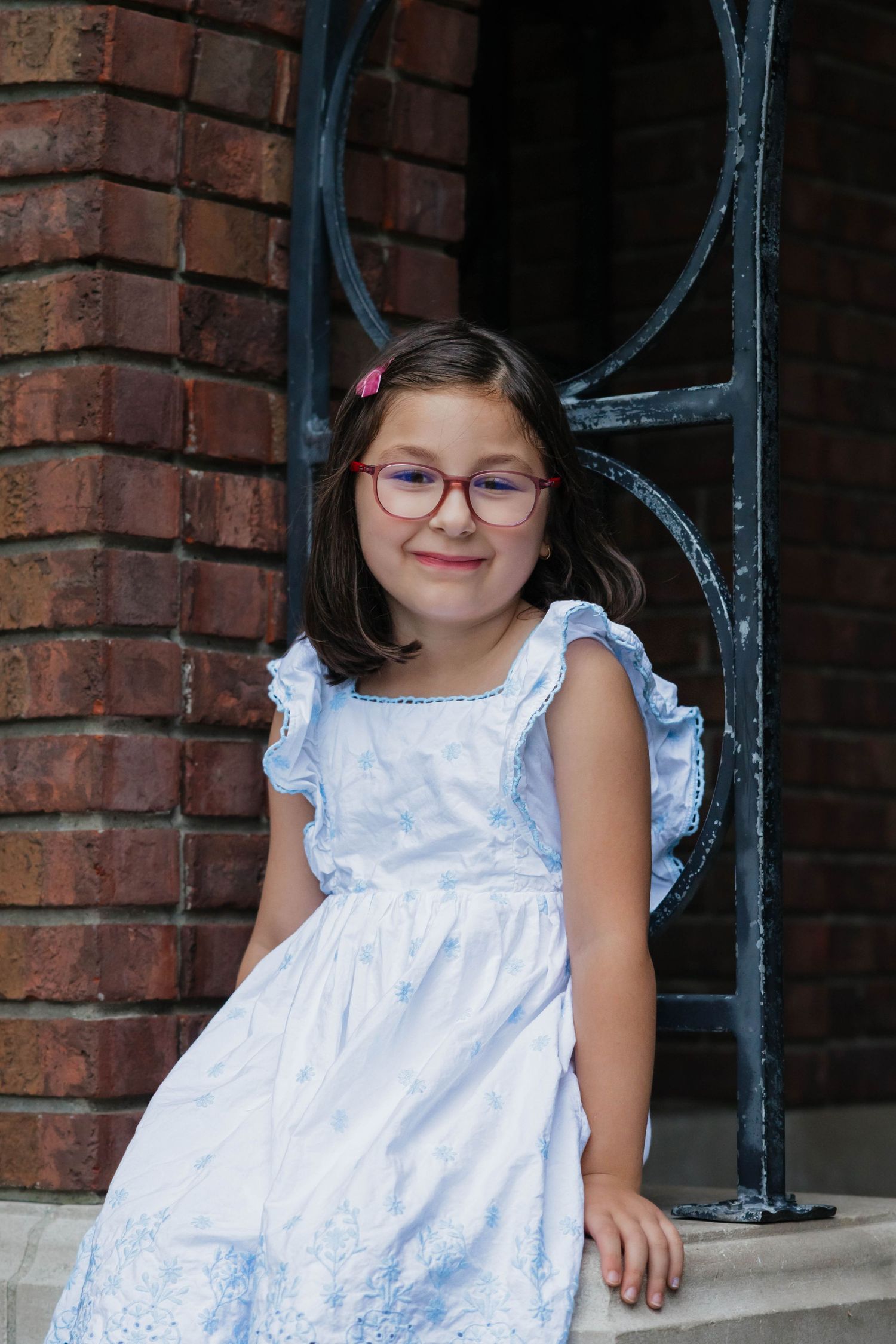 Young girl in a blue floral dress with red glasses leaning against ornate ironwork on a brick building.