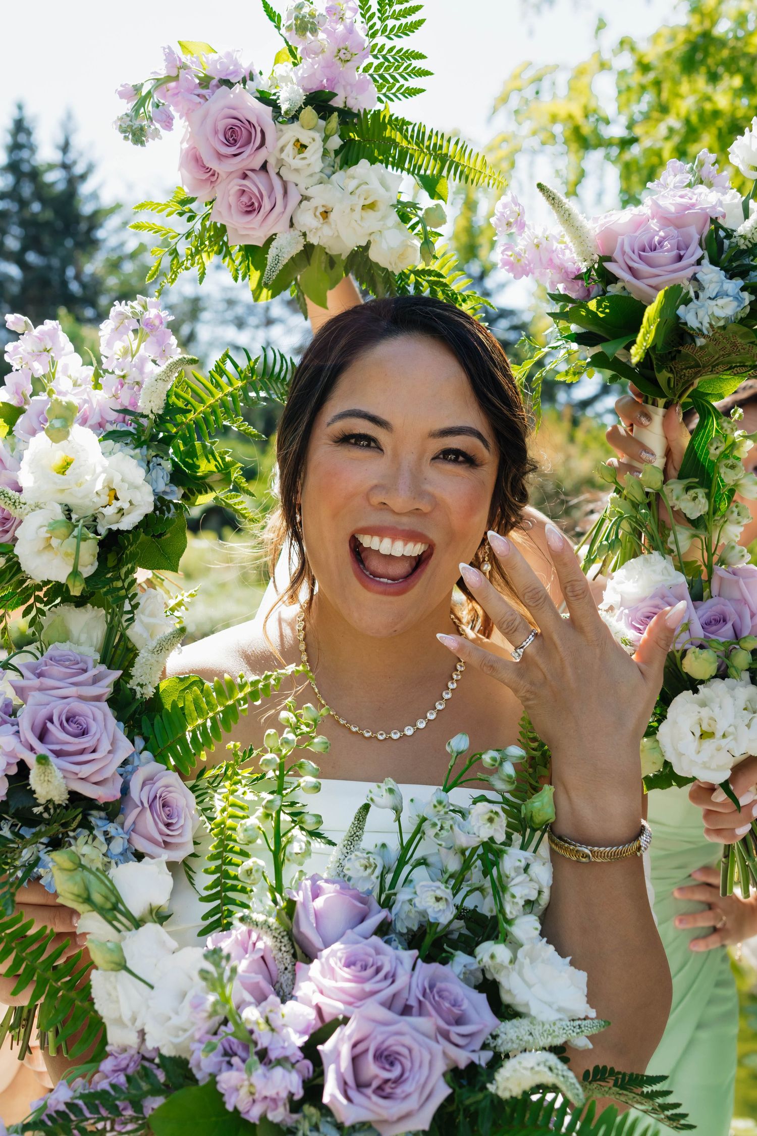Smiling bride shows off wedding ring surrounded by bridesmaids holding lavender and white rose bouquets outdoors.