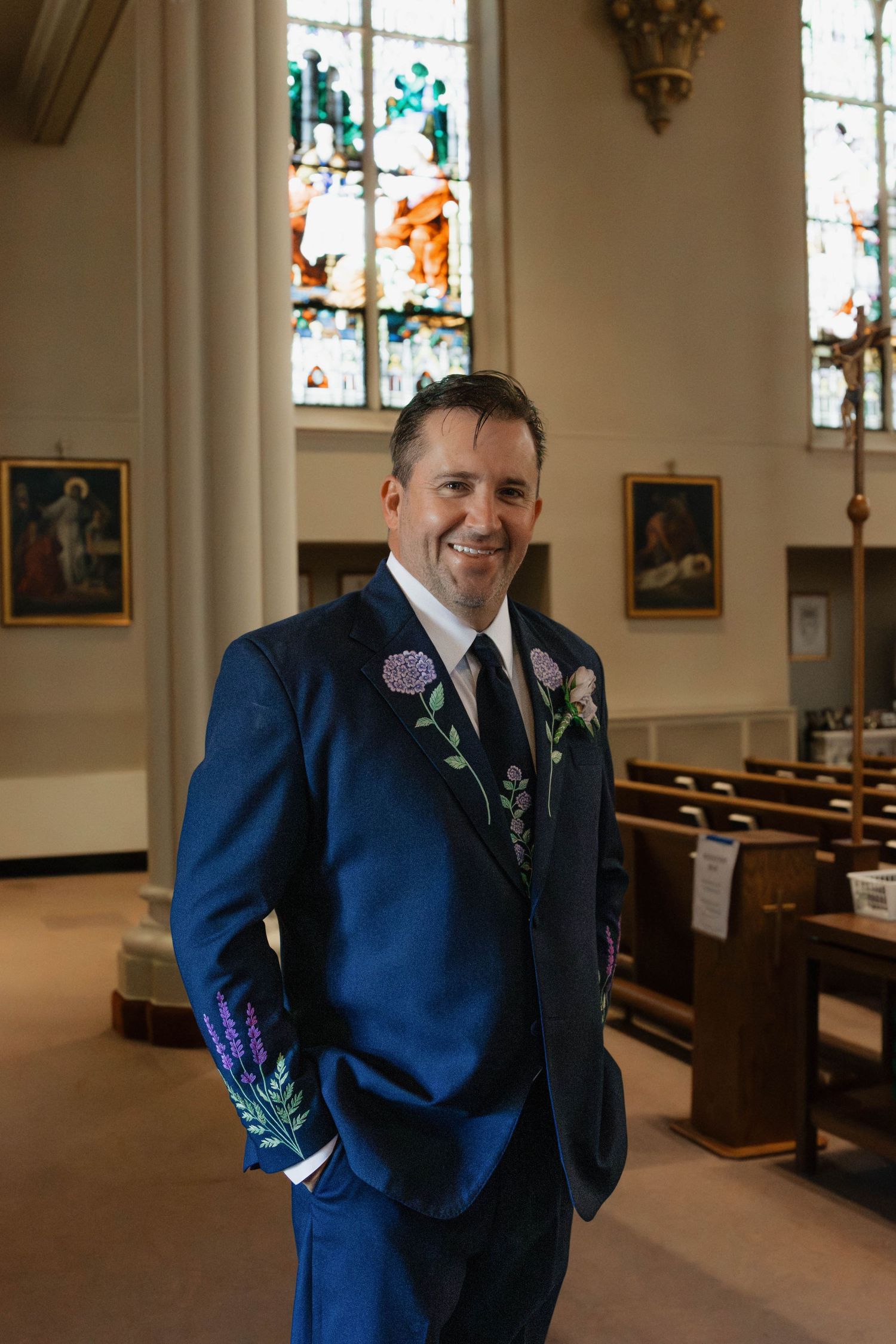 Groom in navy blue floral embroidered suit smiles inside a church with stained glass windows.