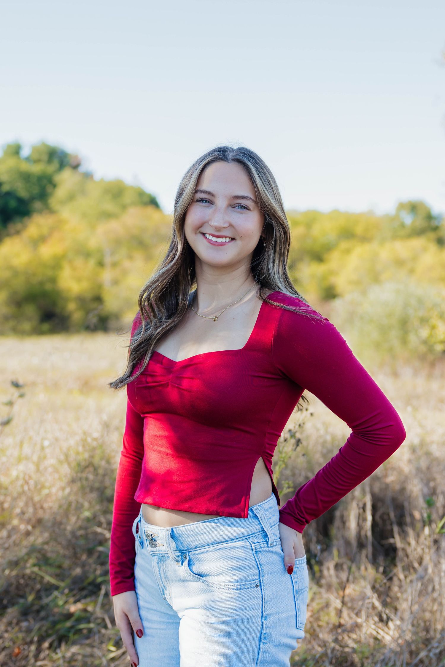 Smiling teenage girl in a red crop top and jeans posing in a sunny autumn field.