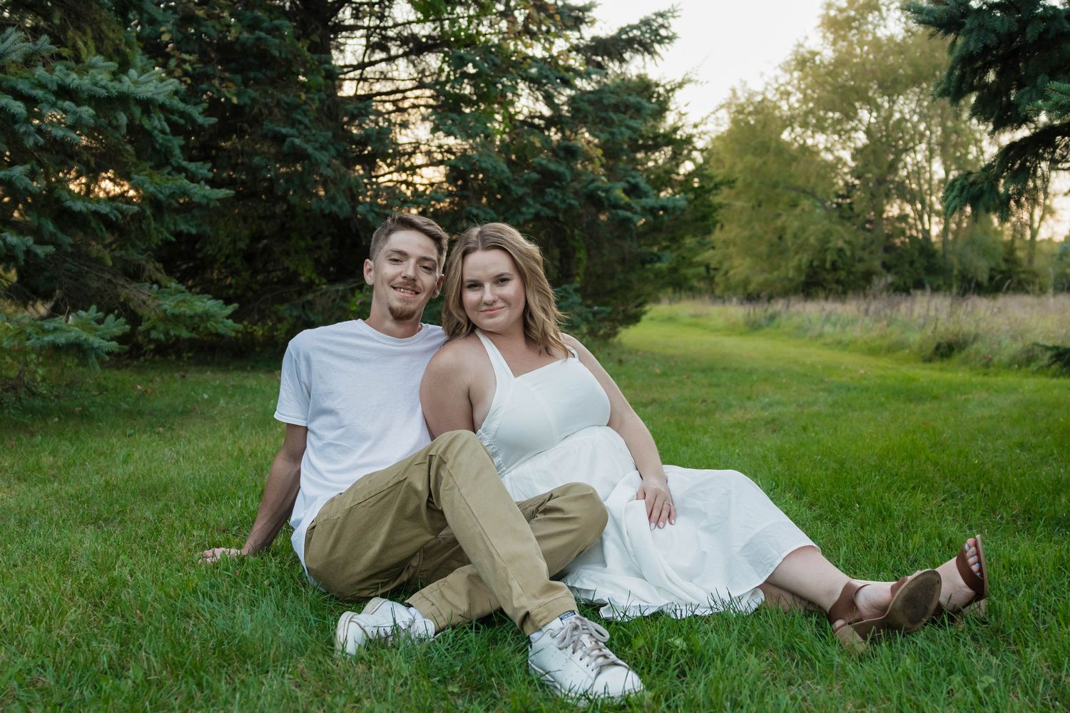 Happy engaged couple sitting on green grass in a park at sunset, smiling at the camera.