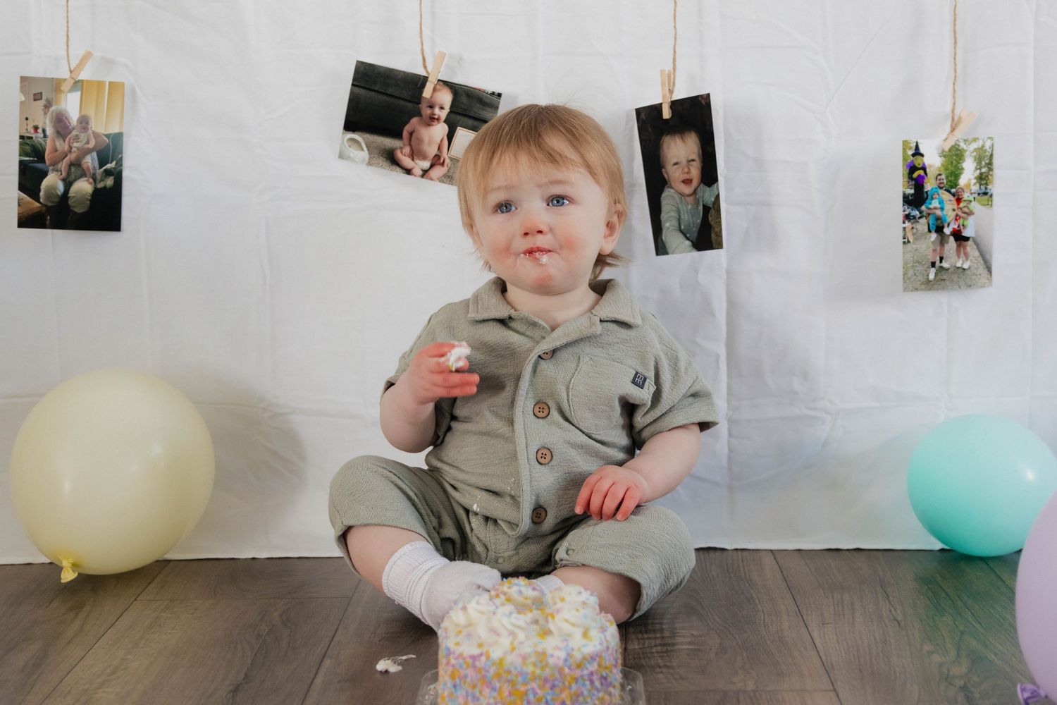 One-year-old boy sits with smashed birthday cake surrounded by colorful balloons on floor.