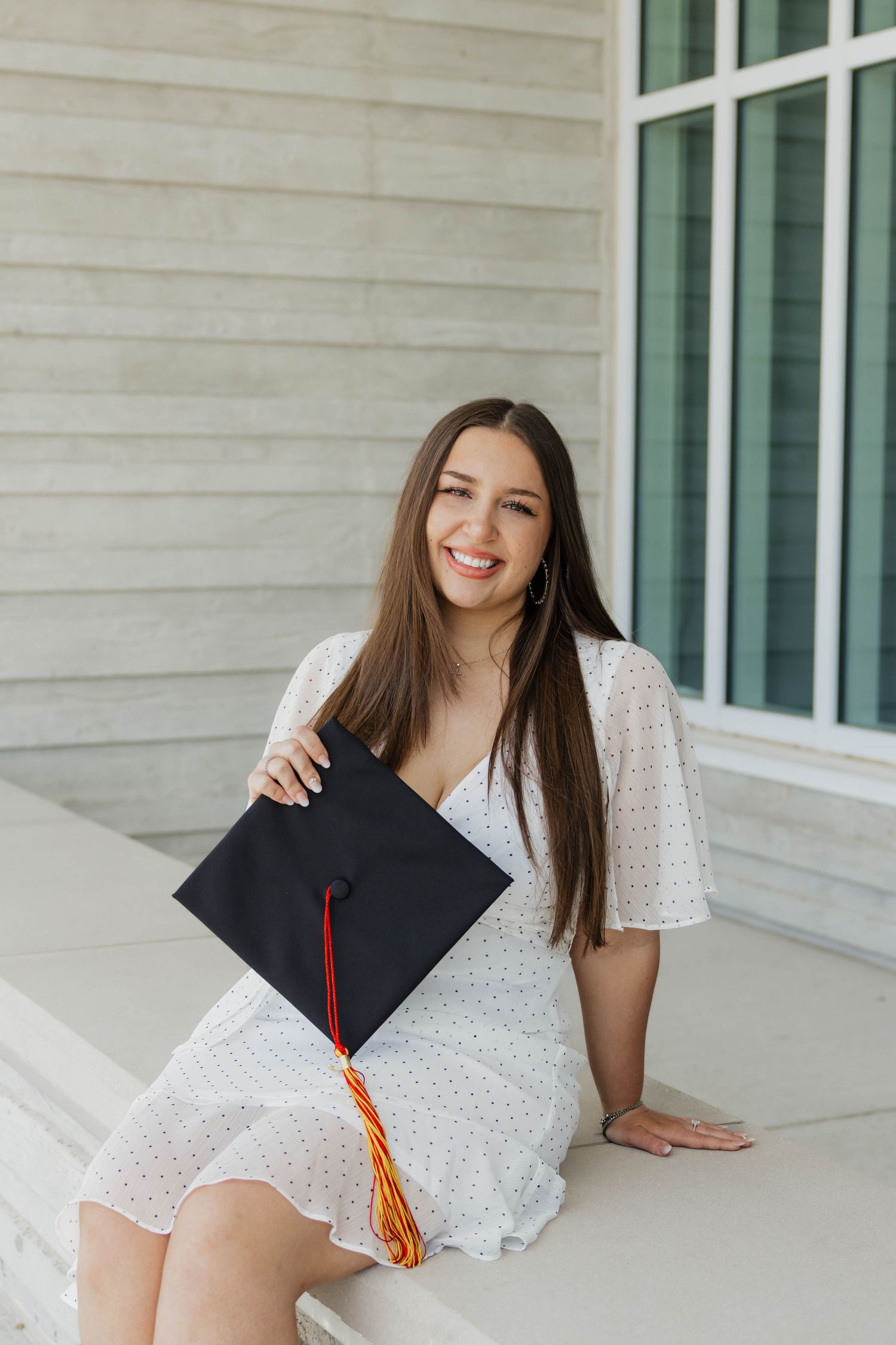 Smiling female graduate in white dress holding black mortarboard cap on university steps.