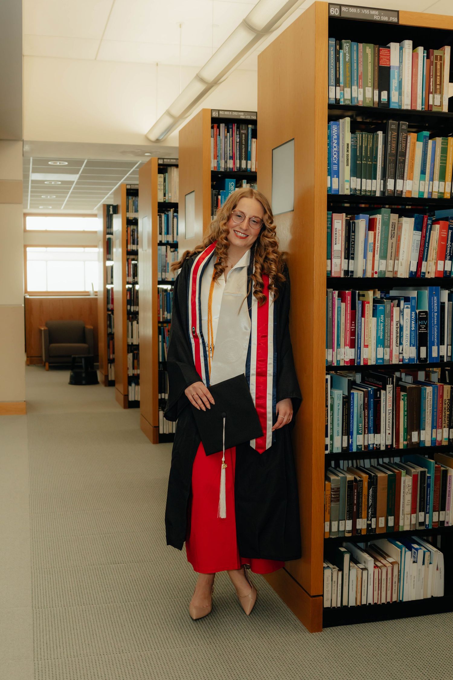 A smiling graduate in a black gown and red dress poses beside library bookshelves holding her cap.