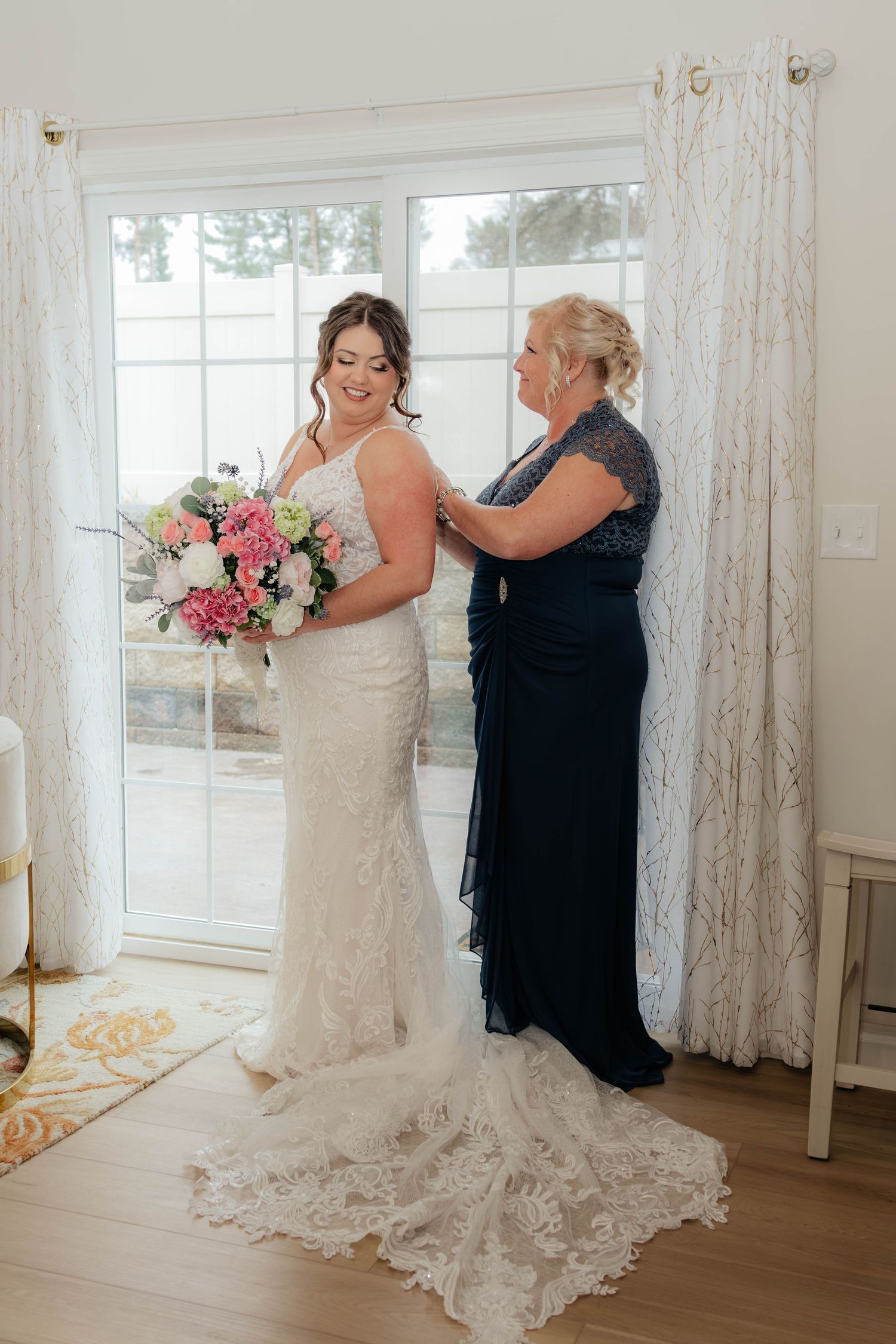 Bride in lace gown holding pink bouquet while older woman in navy dress adjusts her dress.