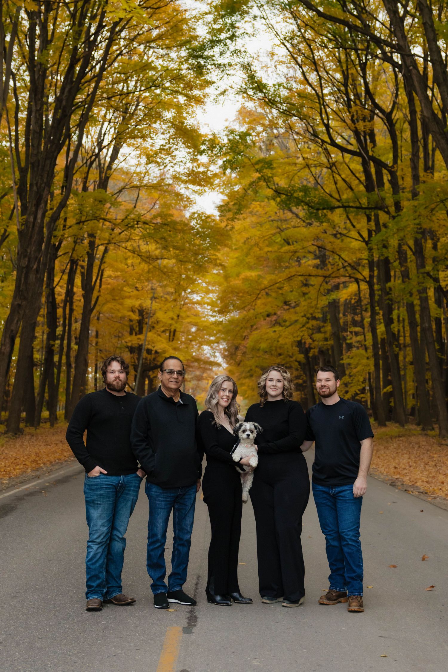 Family of five with small dog posing on tree-lined road surrounded by golden fall foliage.