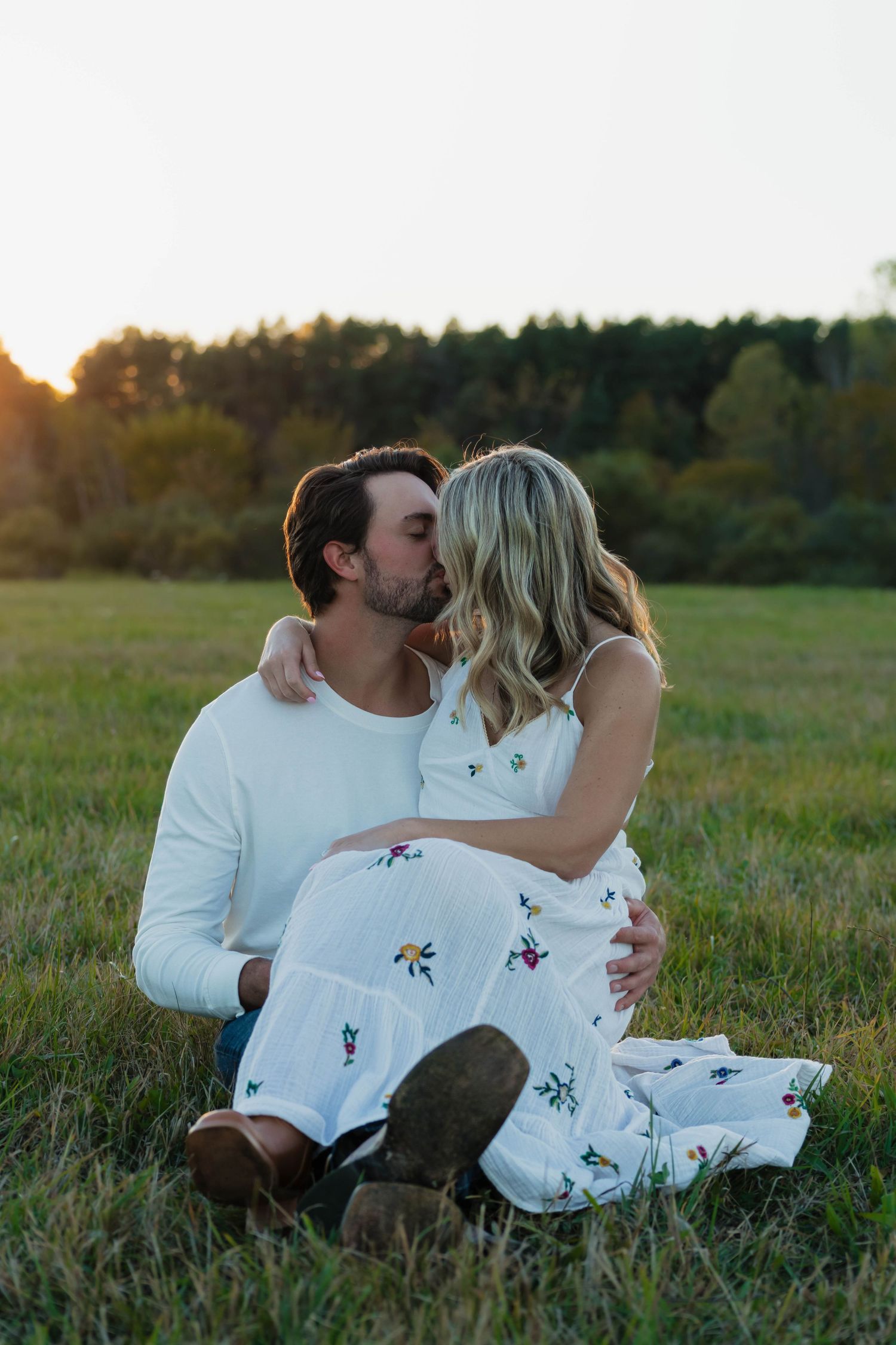 Engaged couple sharing a kiss while sitting in a green meadow during a golden sunset photo session.