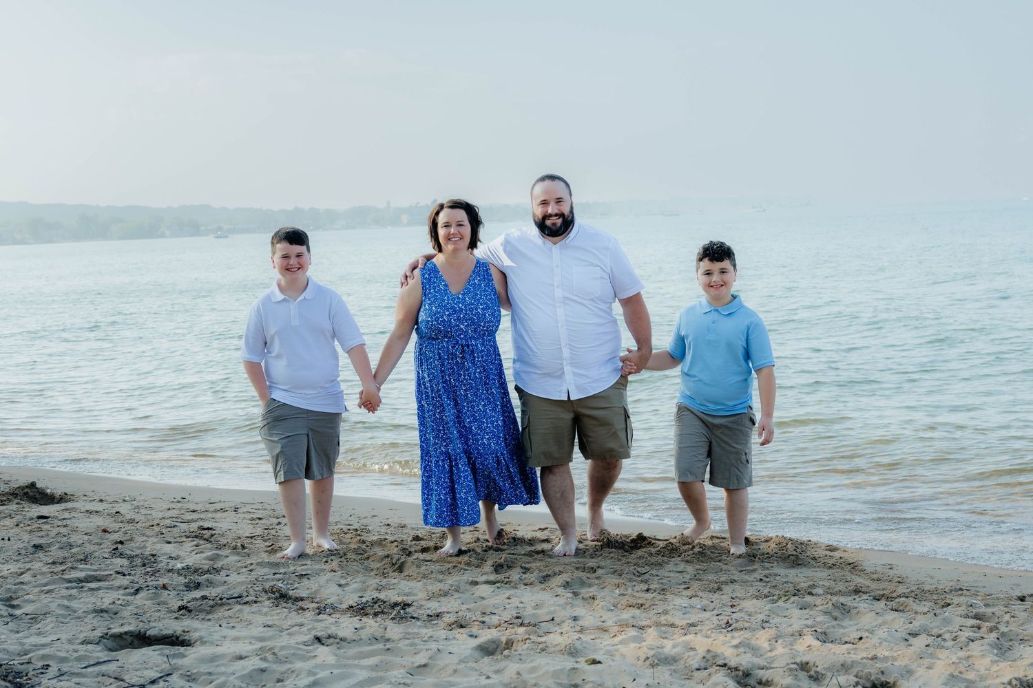 Family of four holding hands and smiling on a sandy beach by the ocean.