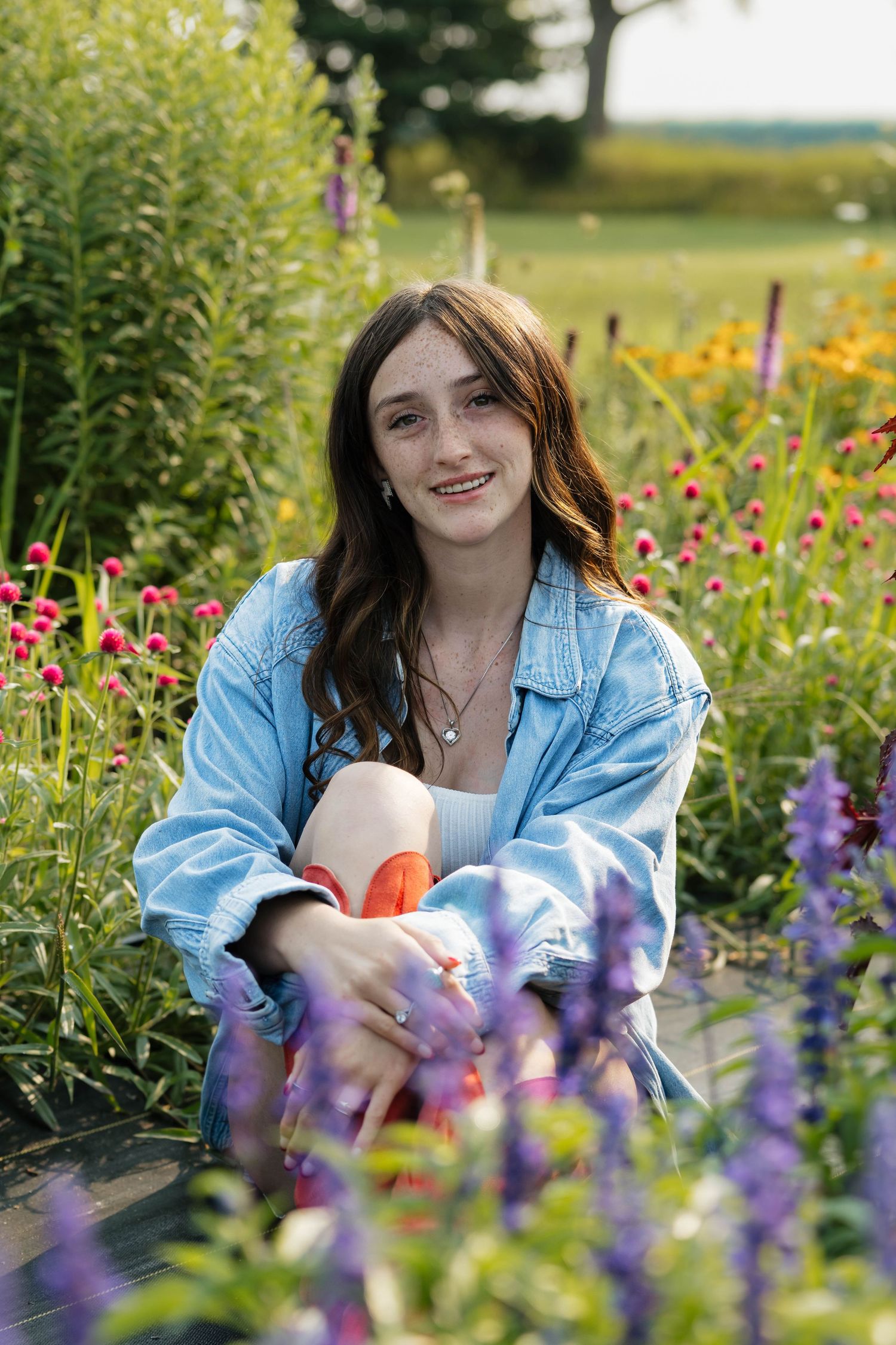Brunette teen smiling in denim jacket among colorful garden flowers.