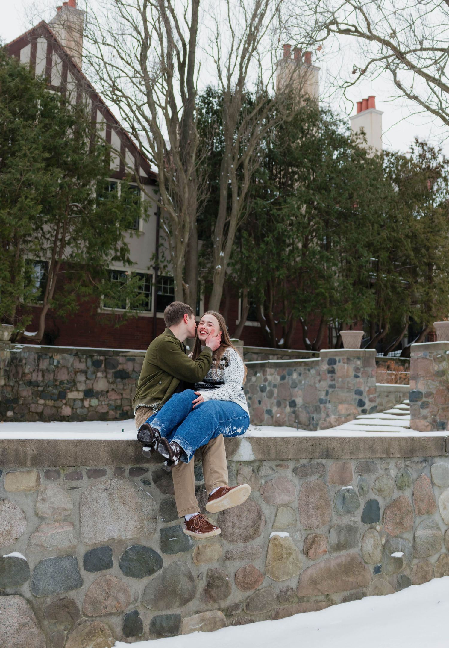 Playful couple sitting on a snowy stone wall, laughing and enjoying a winter engagement session.