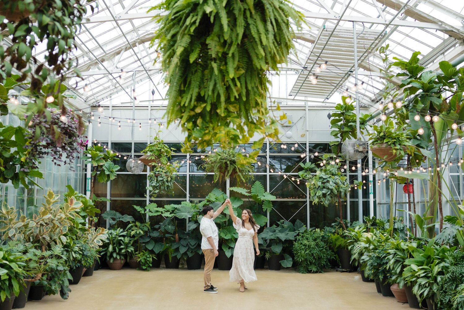 Couple dancing playfully under hanging tropical plants in a bright sunlit botanical greenhouse.