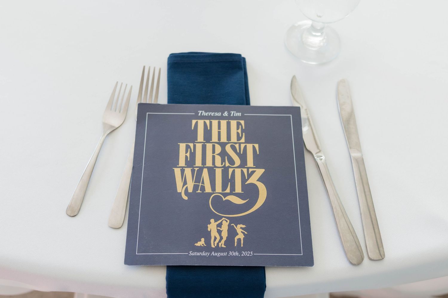 Navy blue wedding program titled The First Waltz placed on white plate with silverware setting.