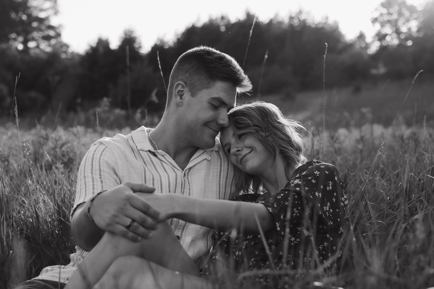 Black and white photo of couple sitting in tall grass, leaning foreheads together and smiling romantically.