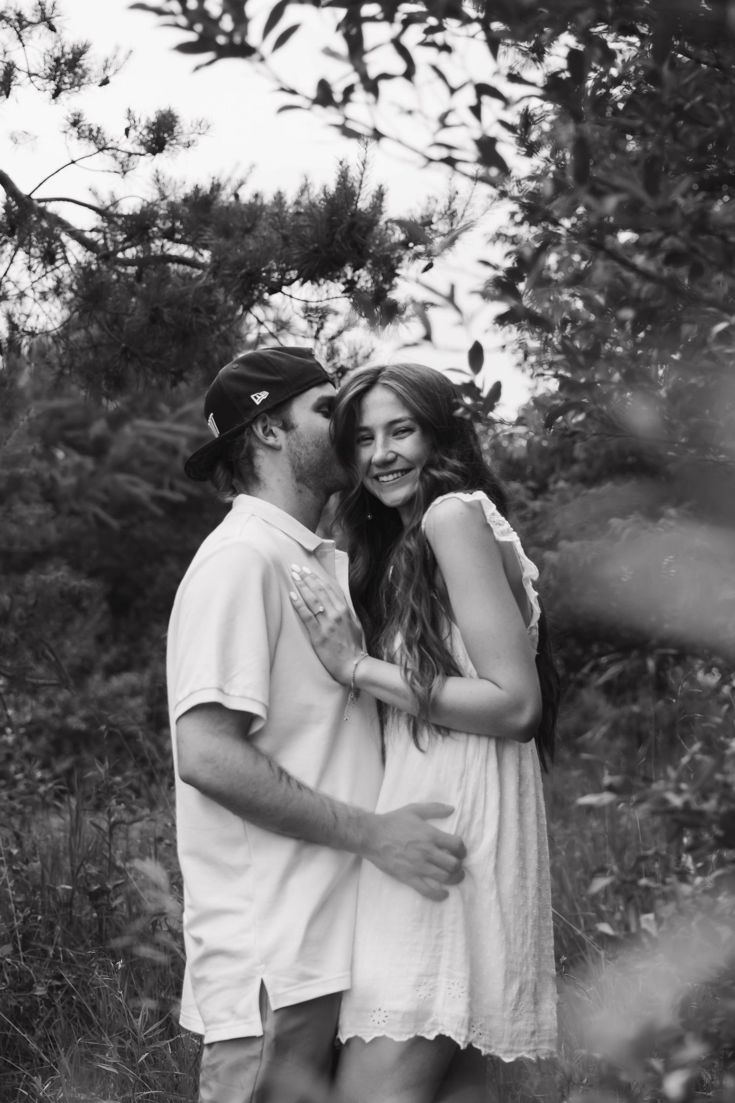 Black and white photo of couple embracing among trees, man kissing woman's cheek as she smiles.
