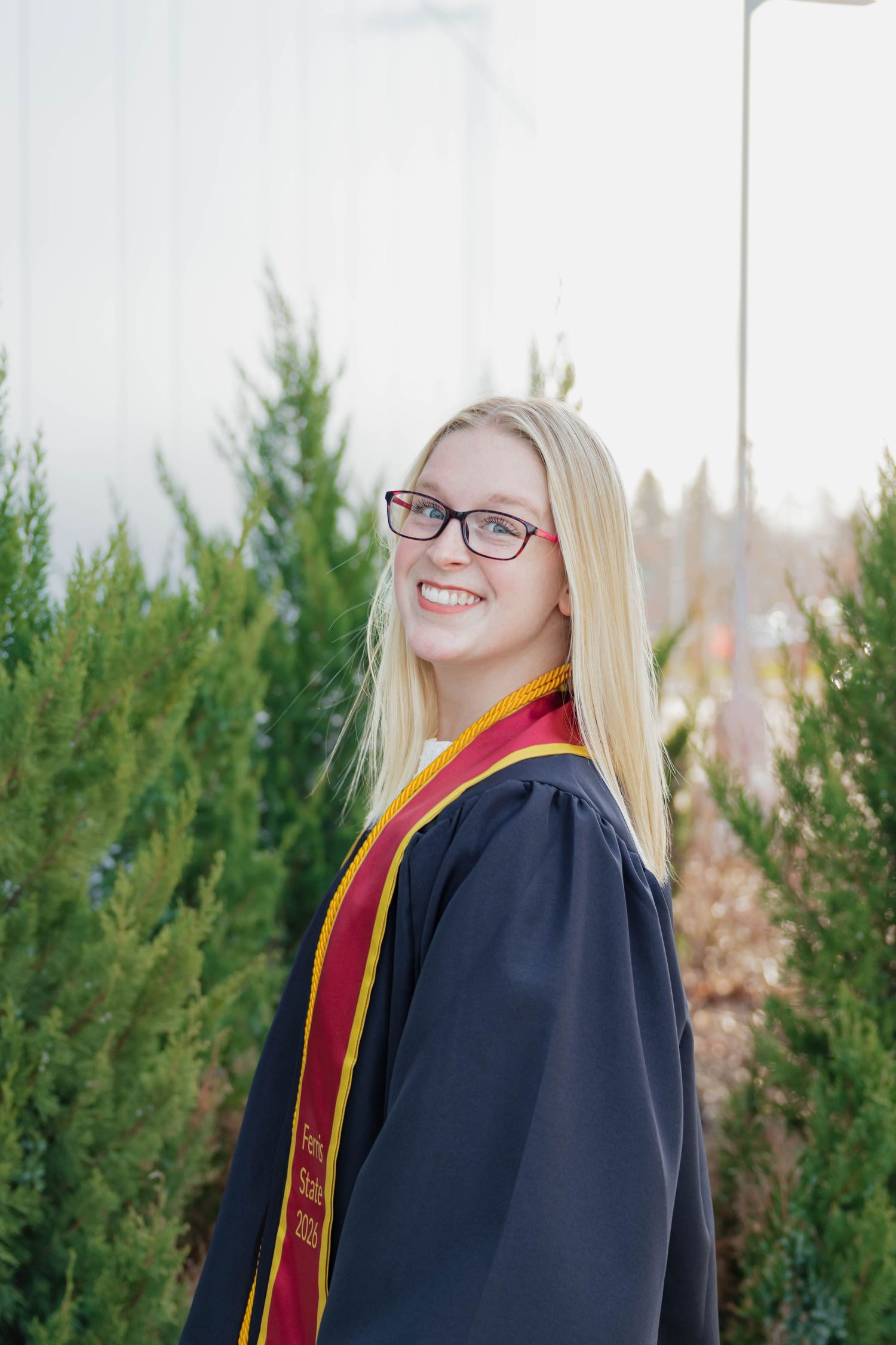 Smiling female graduate in black gown with maroon and gold stole poses outdoors near evergreen trees.