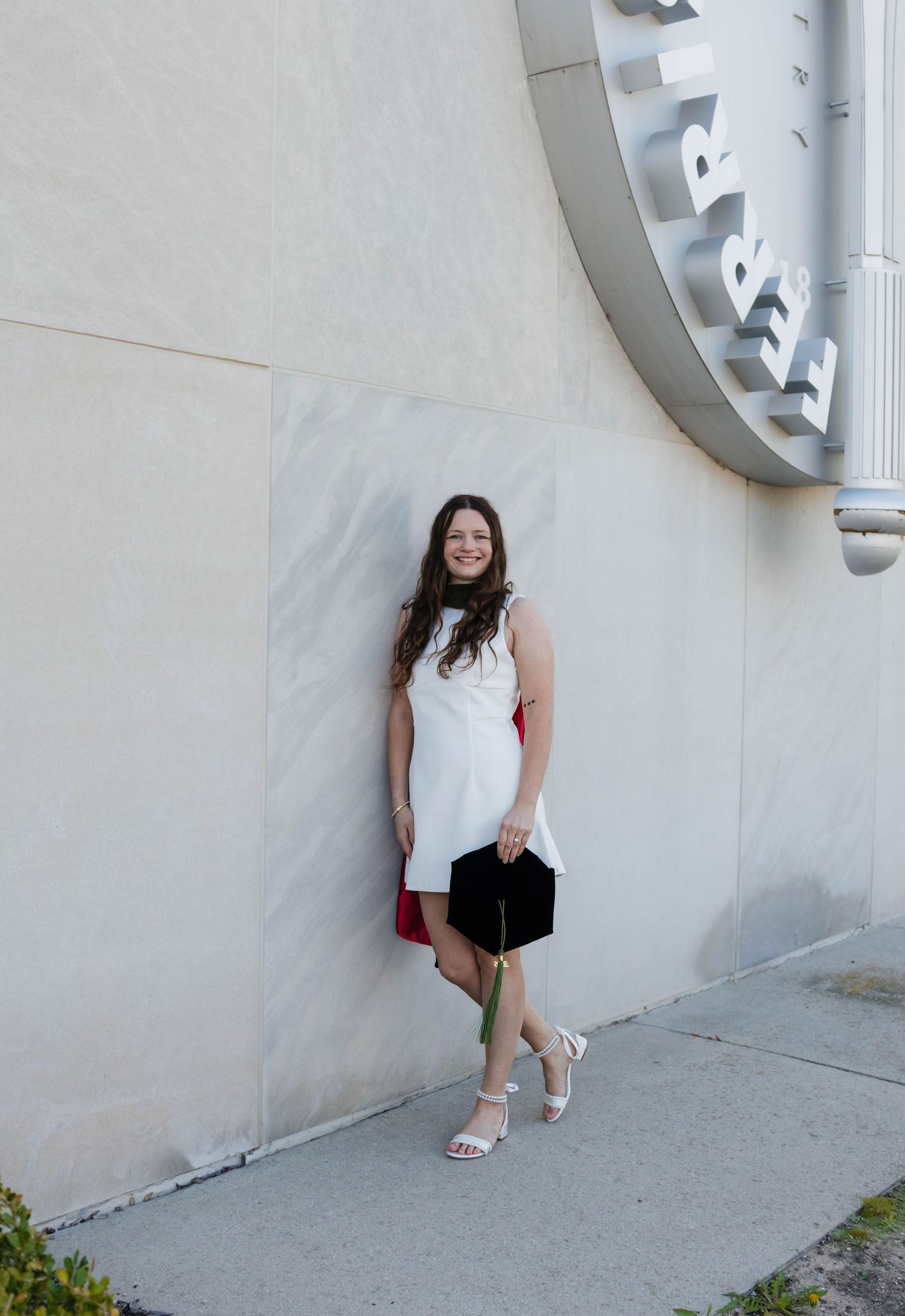Smiling female graduate in white dress leaning against university wall holding graduation cap.