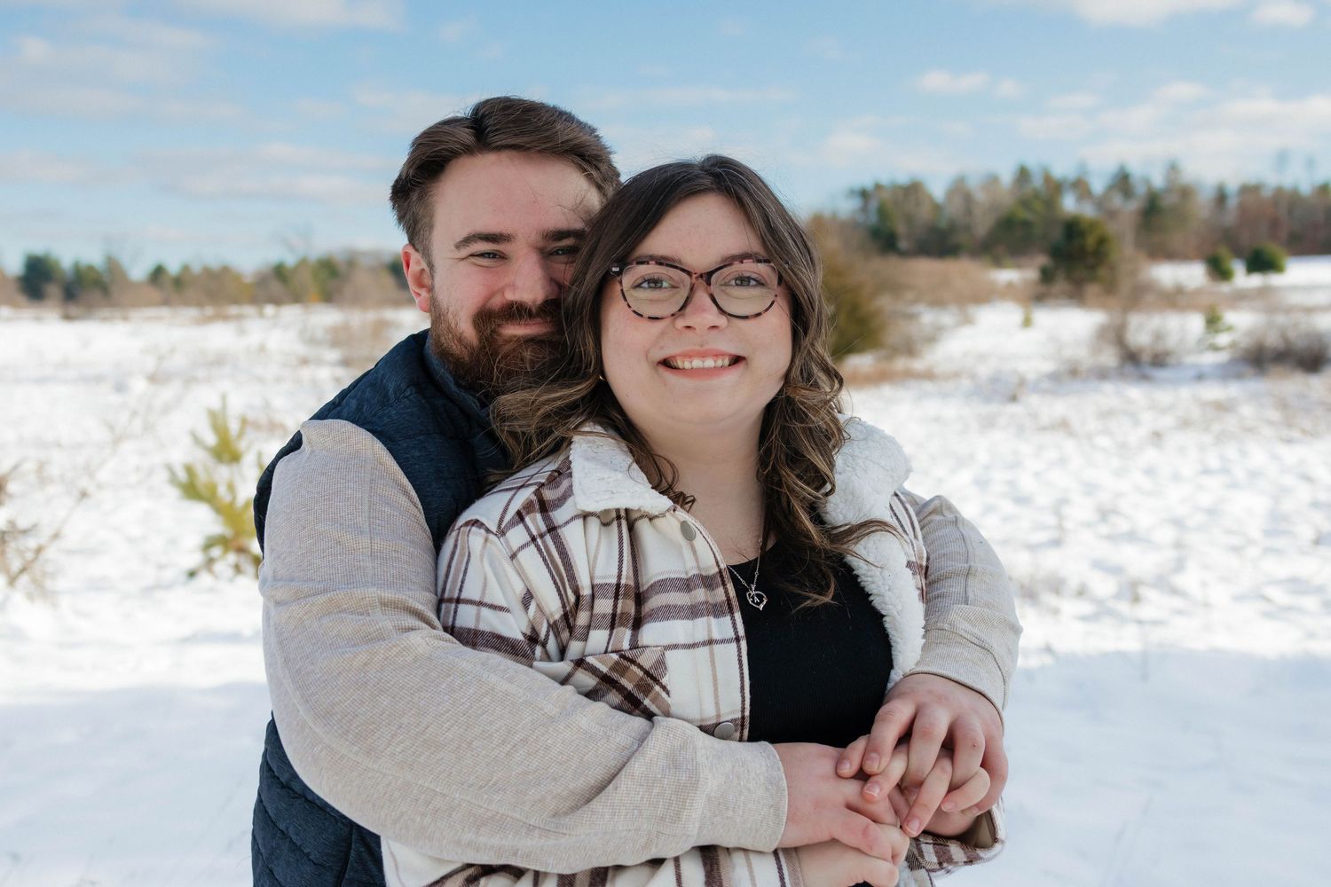 Happy couple hugging and smiling in a snowy winter landscape, woman wearing plaid shacket and glasses.