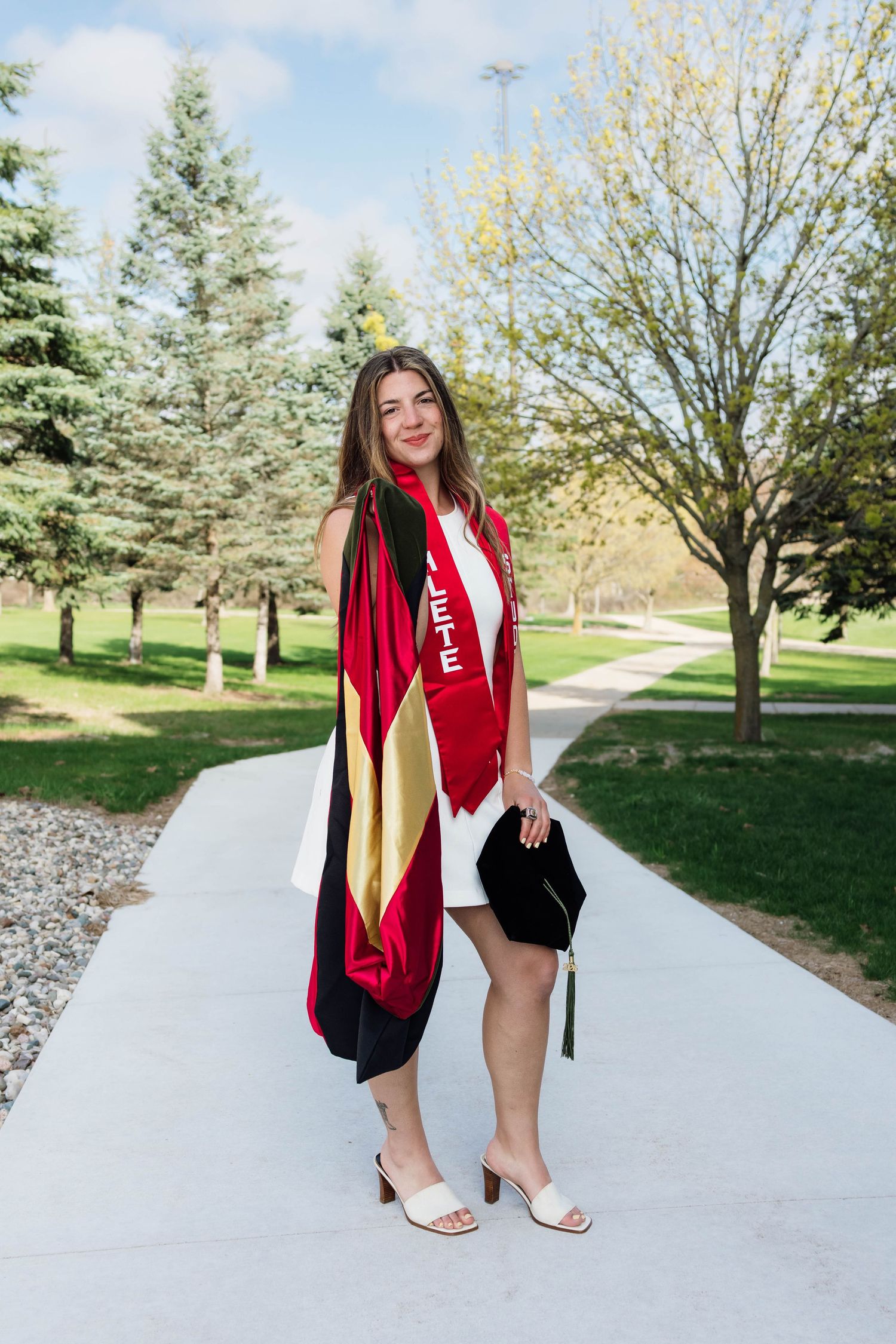 Female graduate in white dress with red sash and graduation cap on tree-lined campus pathway.
