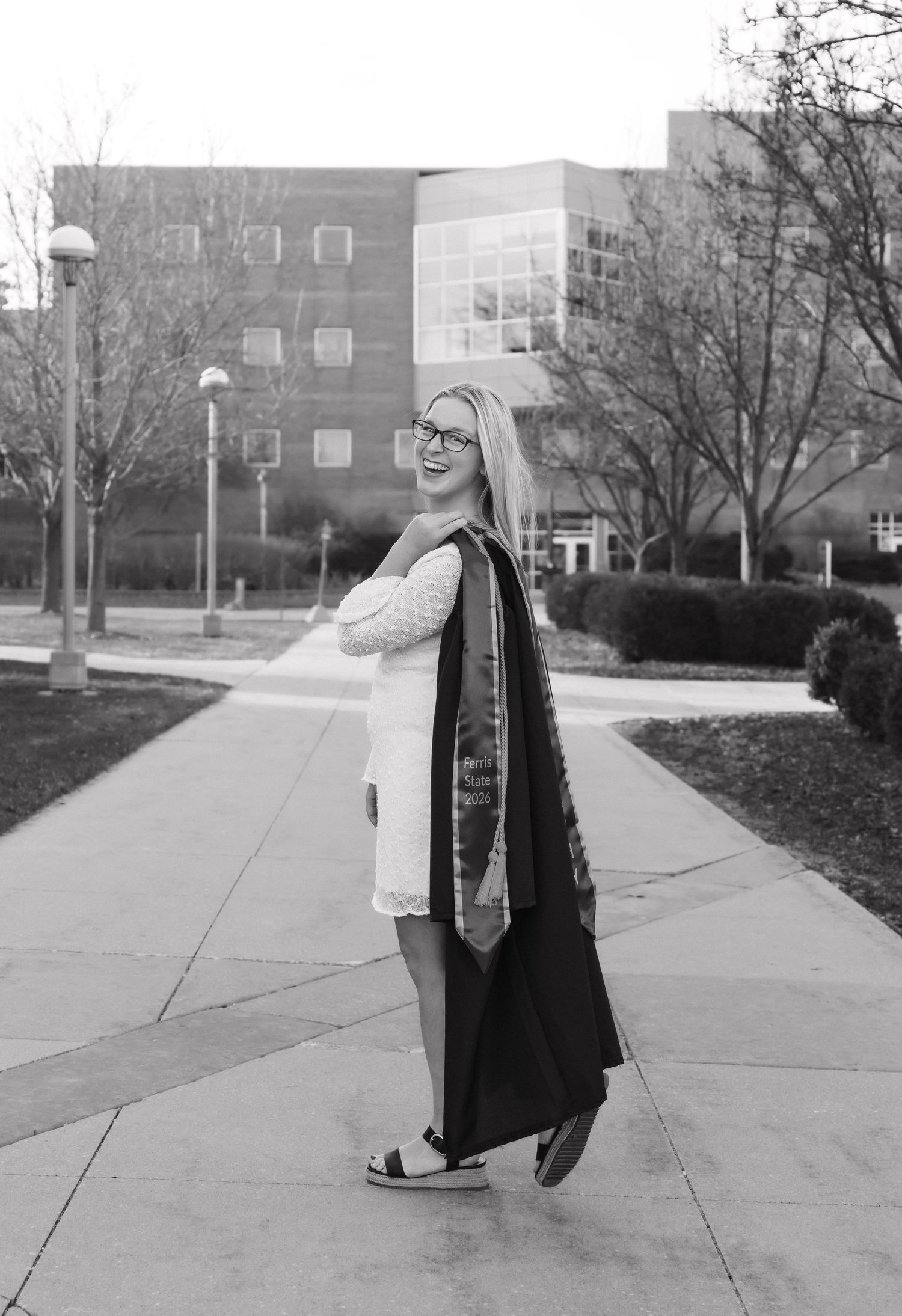 Black and white photo of smiling female graduate in white dress carrying graduation gown on campus path.
