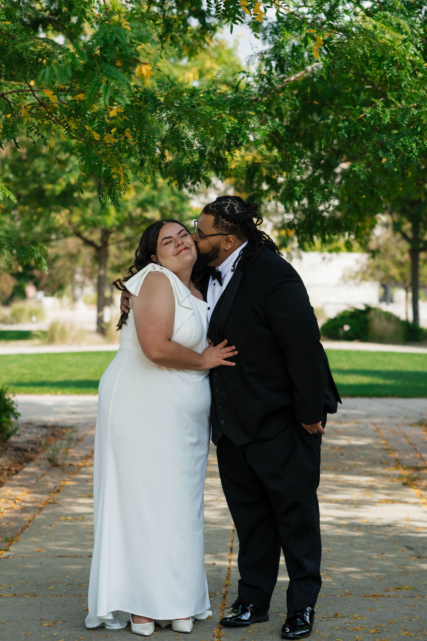 Groom in a black tuxedo kissing his bride in a white gown in a park.