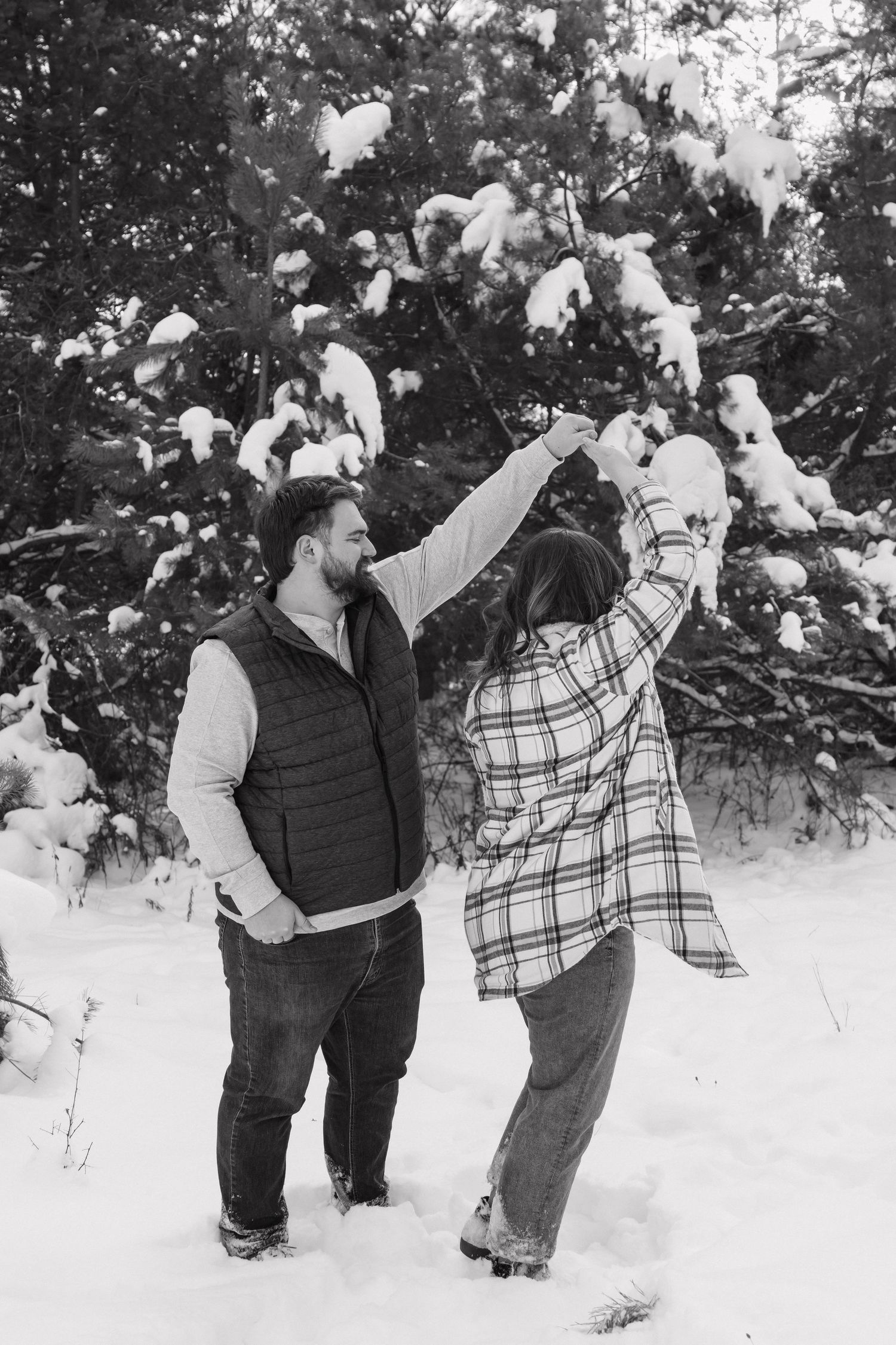 Black and white photo of couple dancing playfully in the snow with snow-laden evergreen trees behind them.