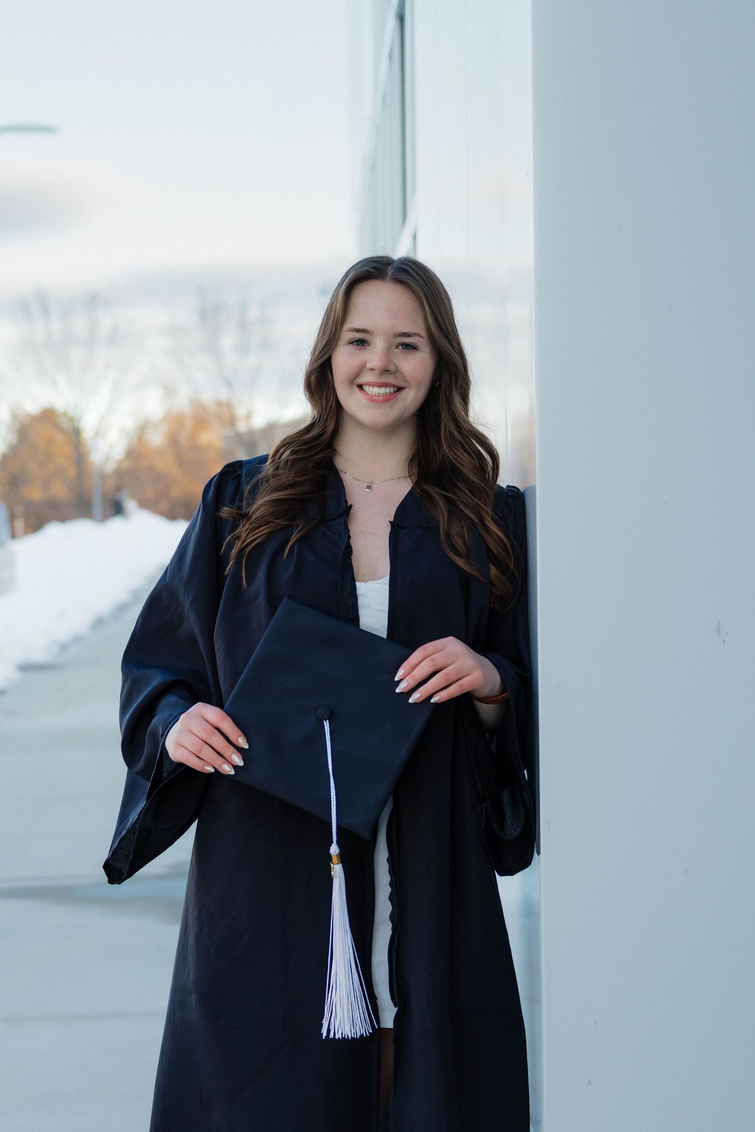 Smiling brunette graduate holding mortarboard cap while leaning against a white pillar outdoors.