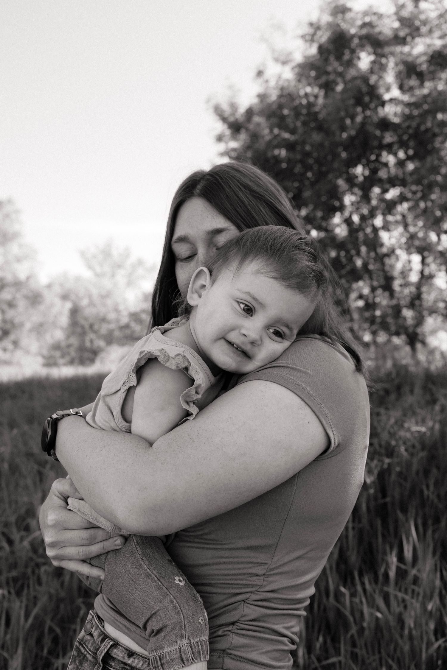 Black and white photo of mother tenderly embracing smiling toddler daughter outdoors in nature.