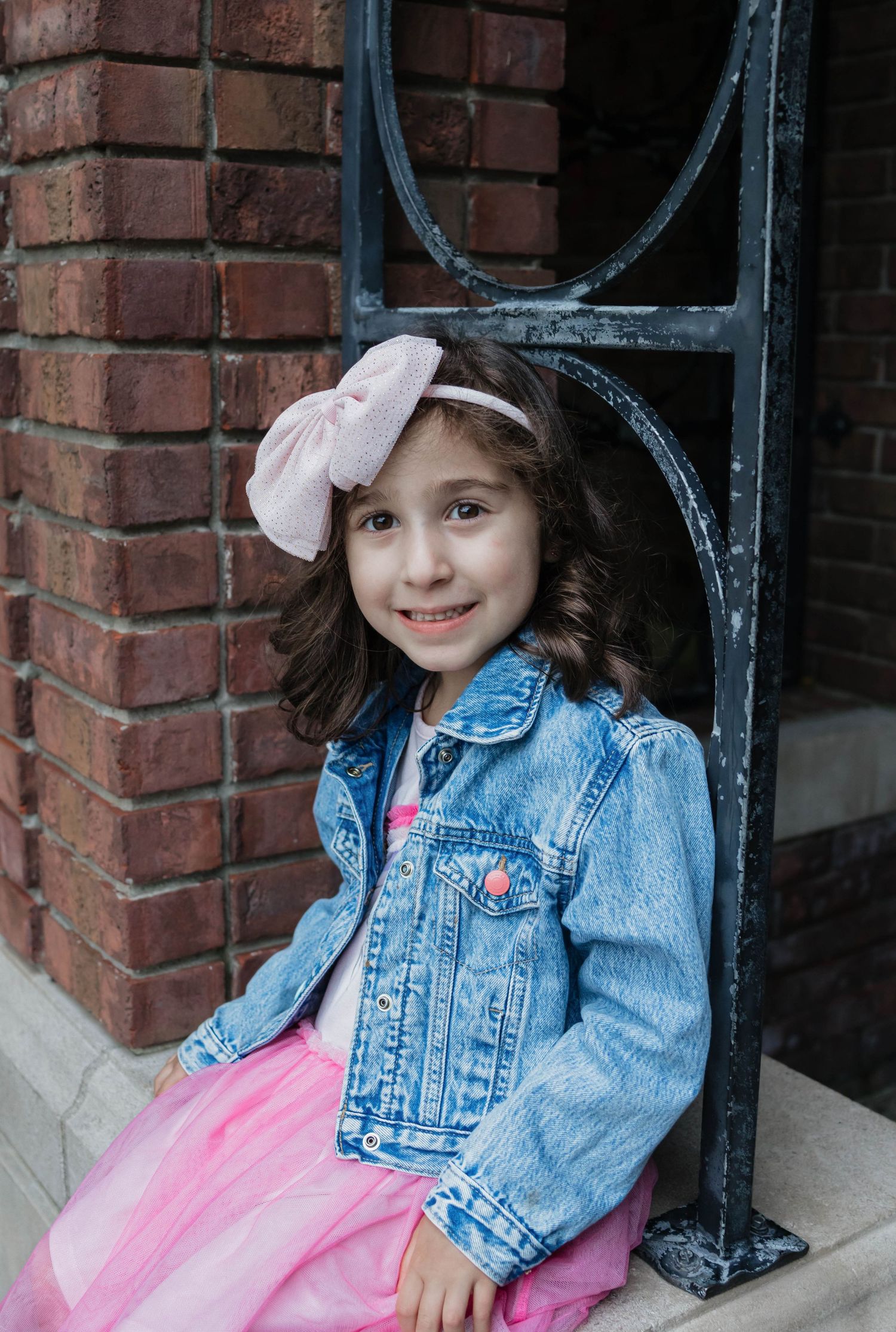 Young girl in denim jacket and pink tutu dress with bow headband poses by brick wall and iron railing.