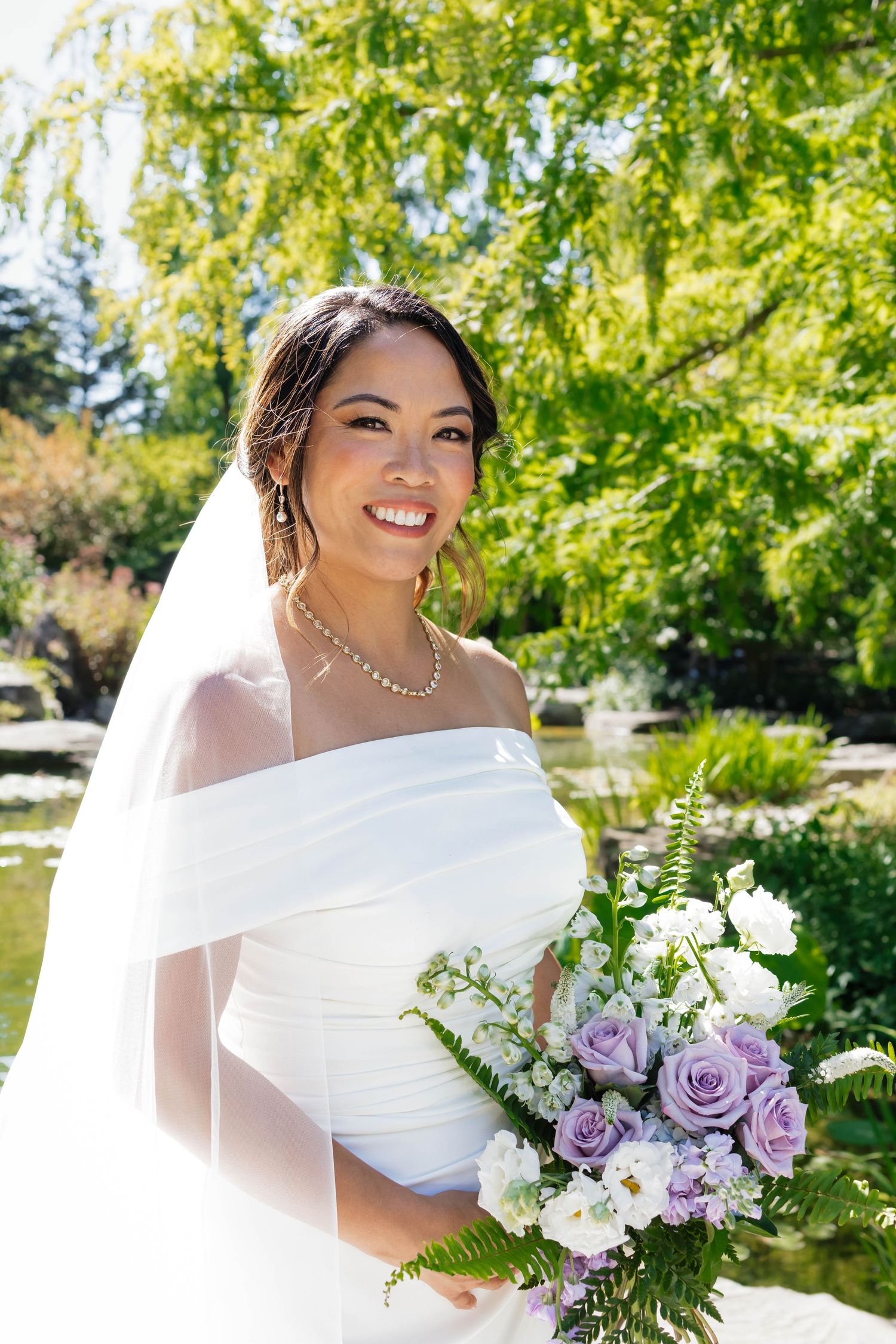 Elegant bride in white off-shoulder gown and veil holds purple and white bouquet in lush garden setting.
