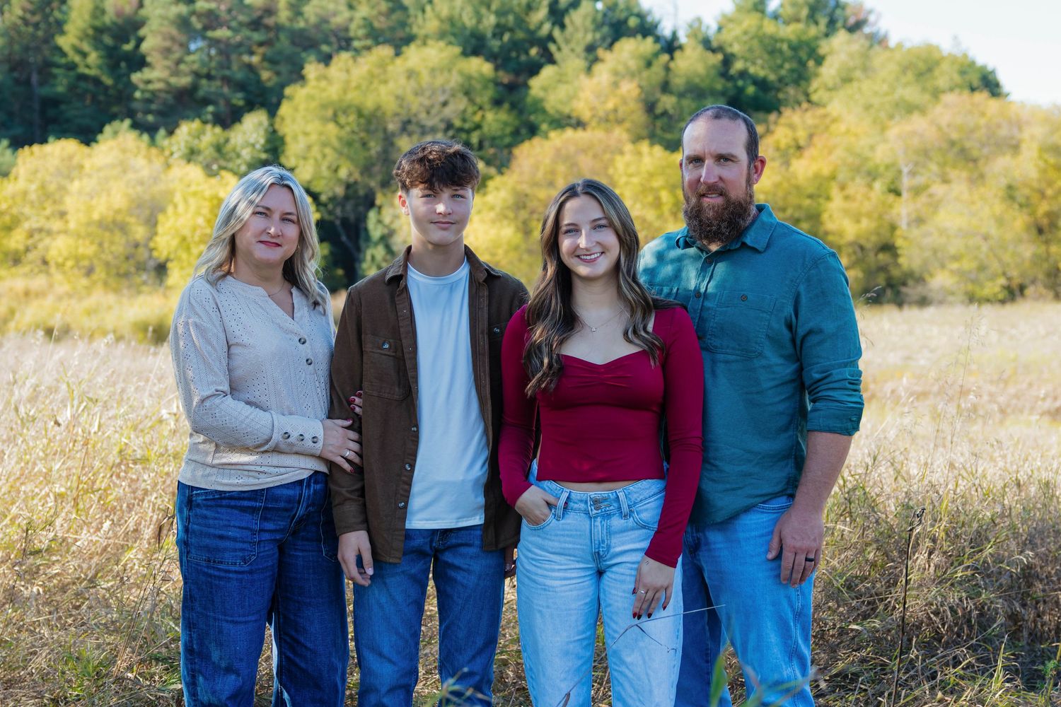 Family of four standing in a golden field with colorful autumn foliage in the background.