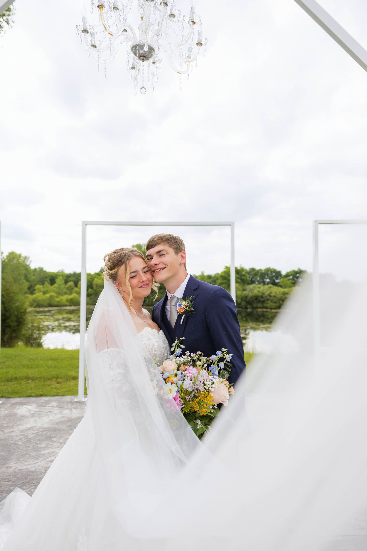 Bride and groom pose romantically through a heart-shaped frame at outdoor wedding ceremony with chandelier overhead.