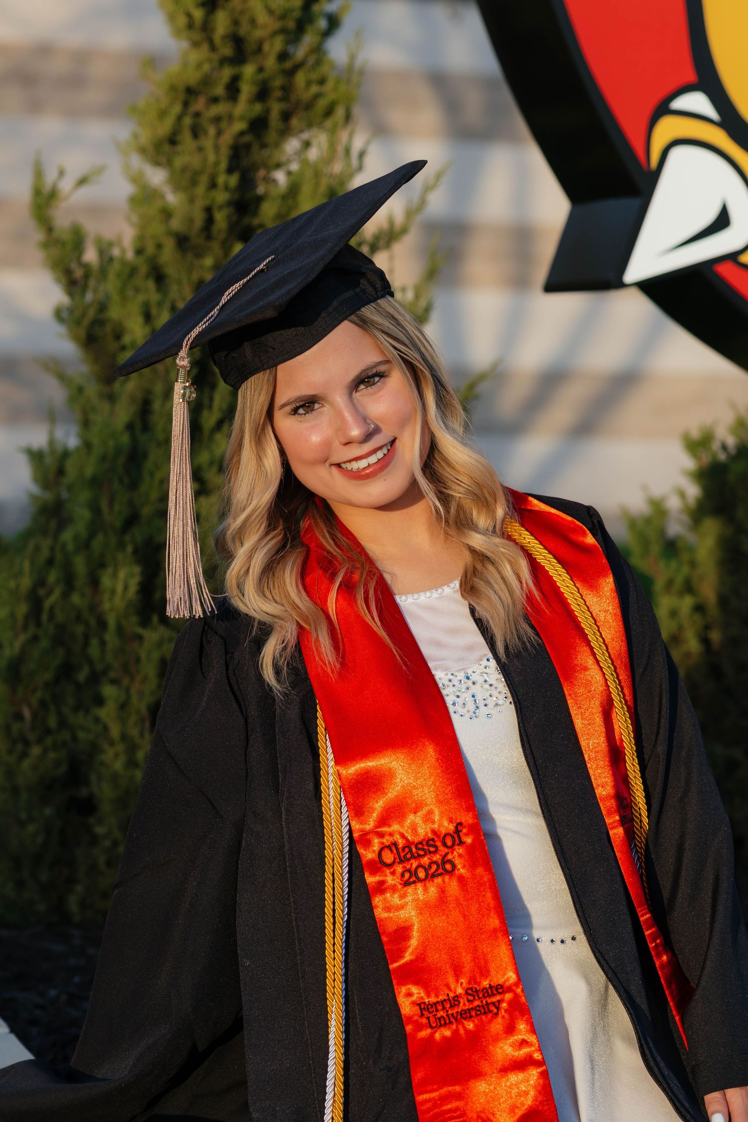Smiling blonde graduate in cap and gown with Ferris State Class of 2025 red satin stole outdoors.
