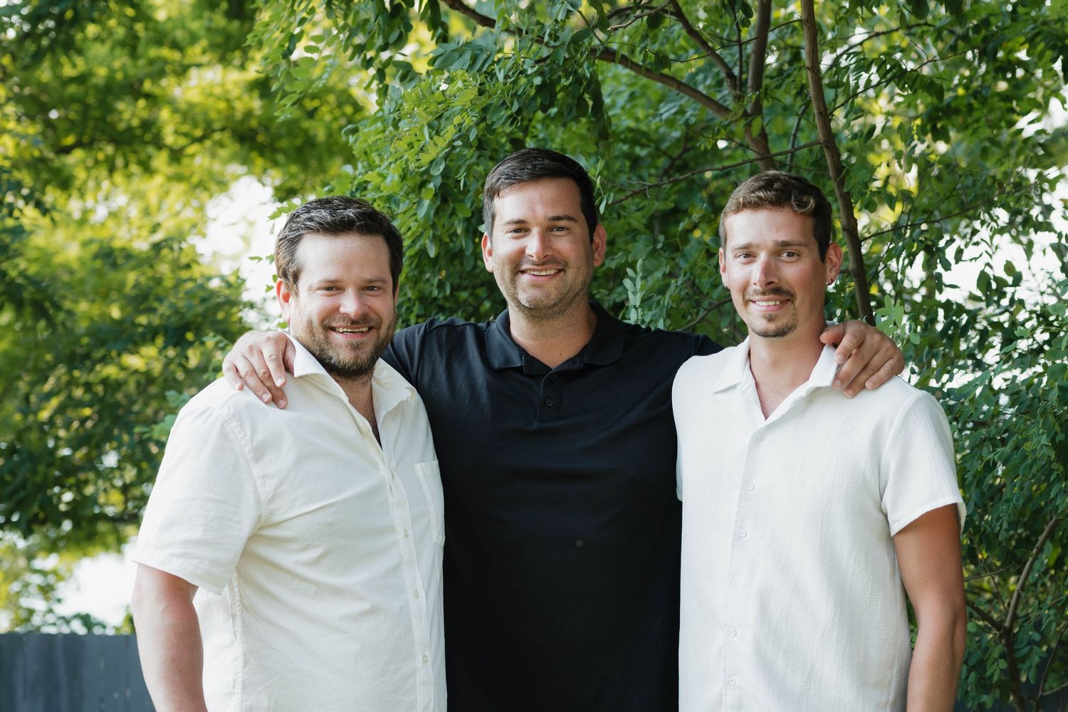 Three smiling adult men standing together outdoors with green trees in the background.