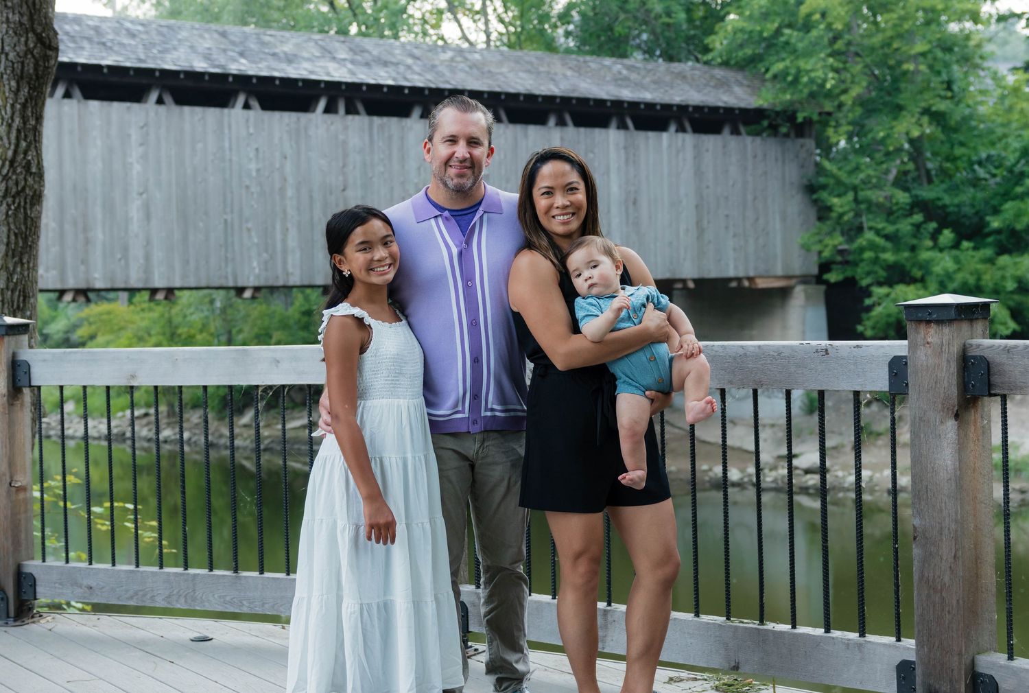 Family of four posing together in front of a wooden covered bridge.