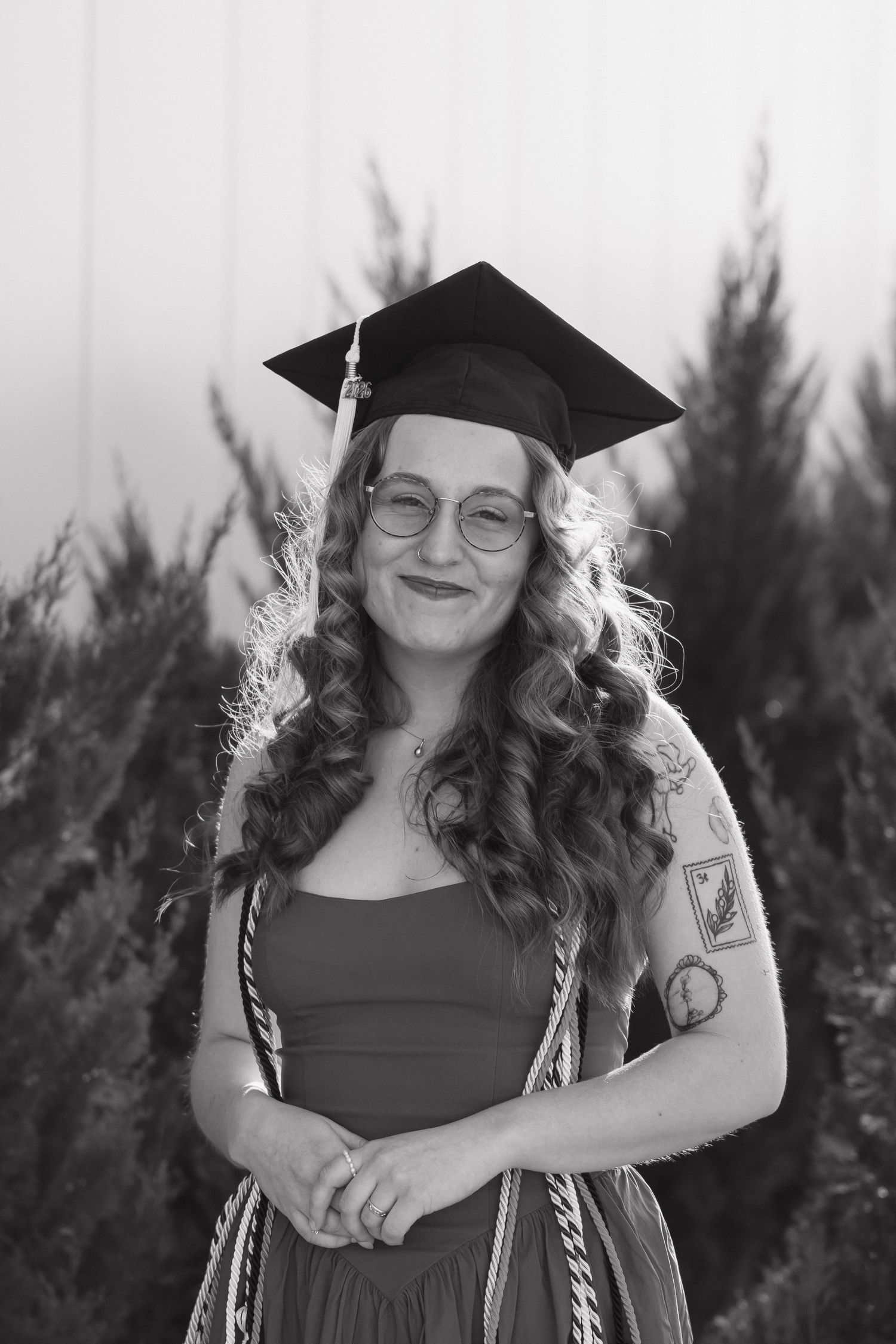Black and white portrait of a smiling graduate in a mortarboard cap and glasses outdoors.