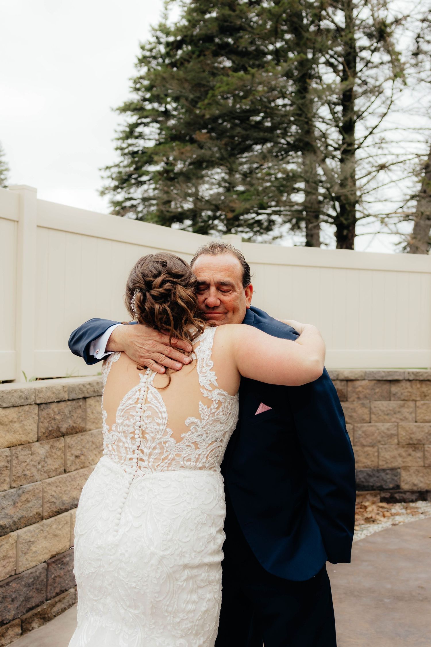 Emotional father hugging bride in lace backless wedding gown outdoors during first look moment.