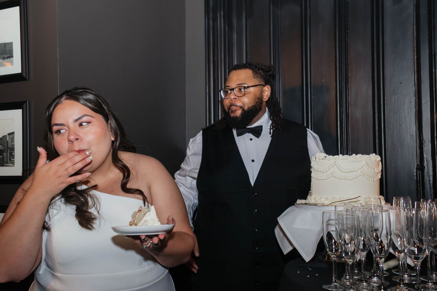 Bride tasting wedding cake while groom holds white tiered cake at reception venue.