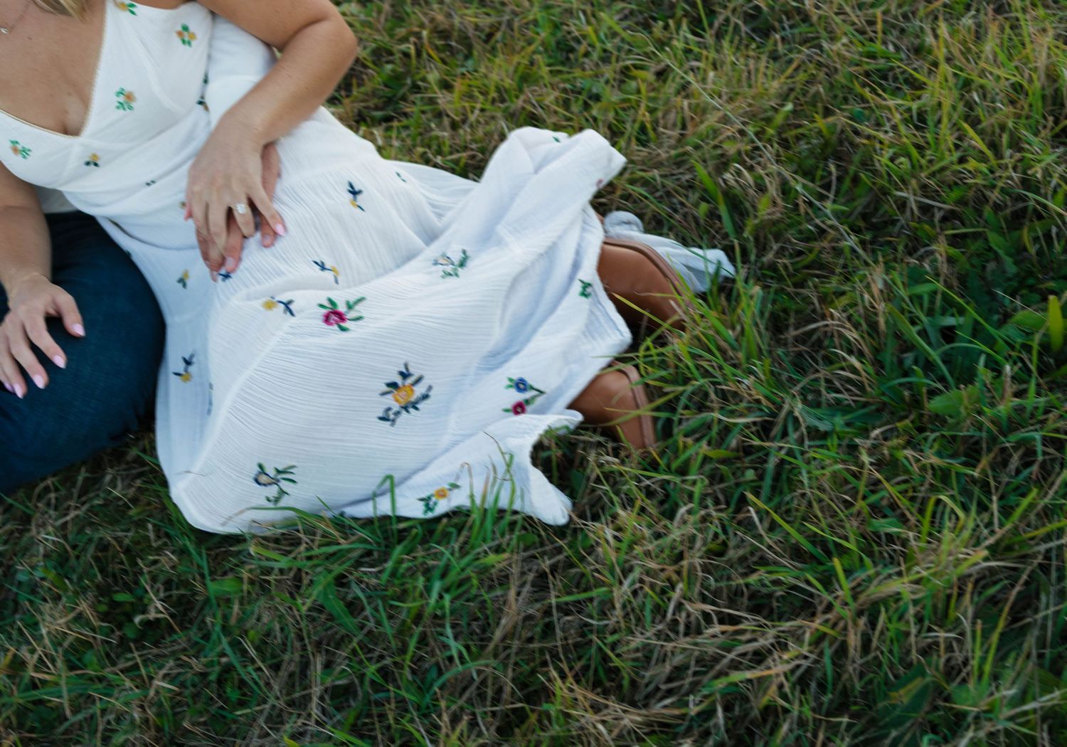 Close-up of a couple sitting in grass, woman wearing a white floral embroidered dress and man in jeans.