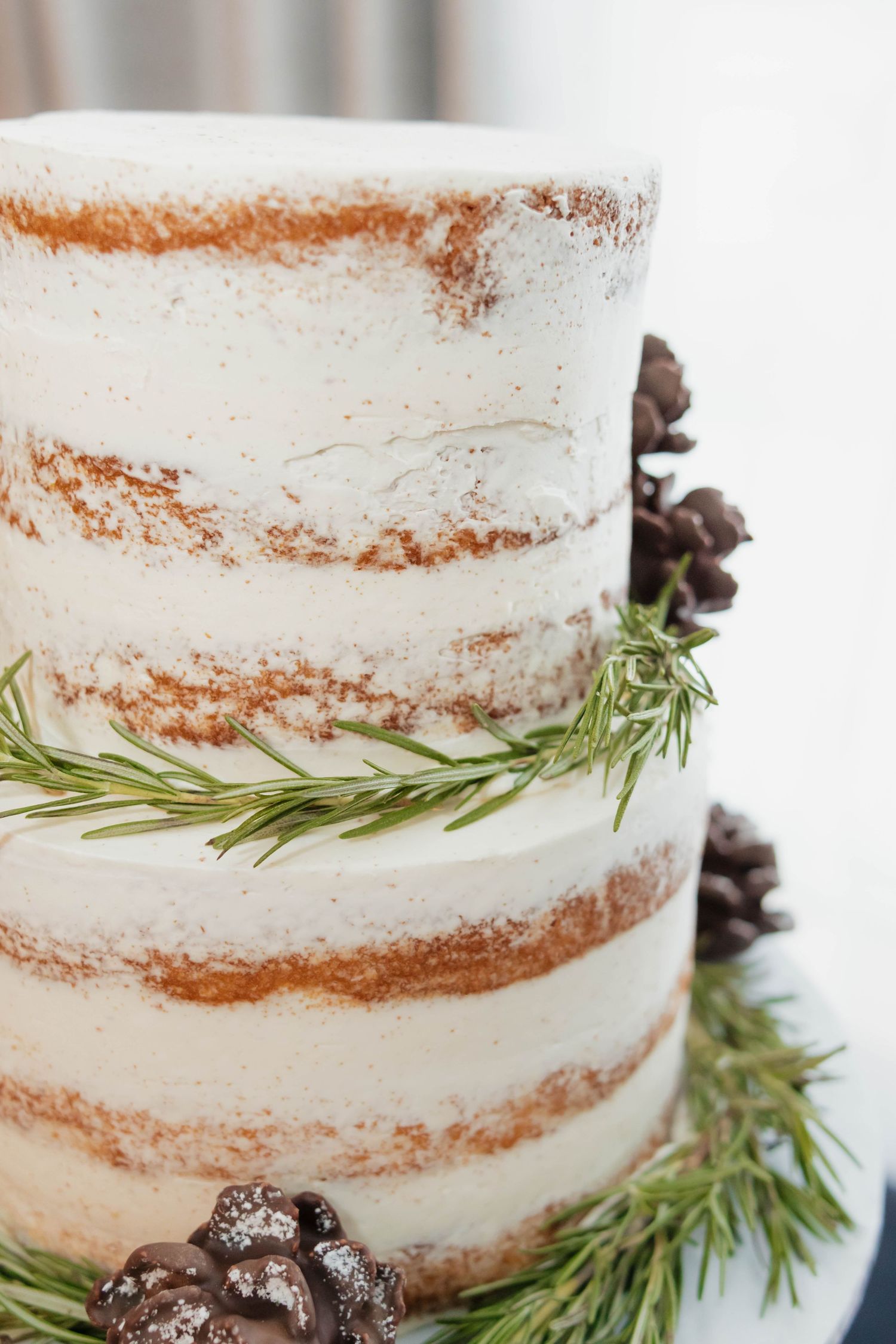 Elegant two-tier naked wedding cake decorated with rosemary sprigs and chocolate pinecones.